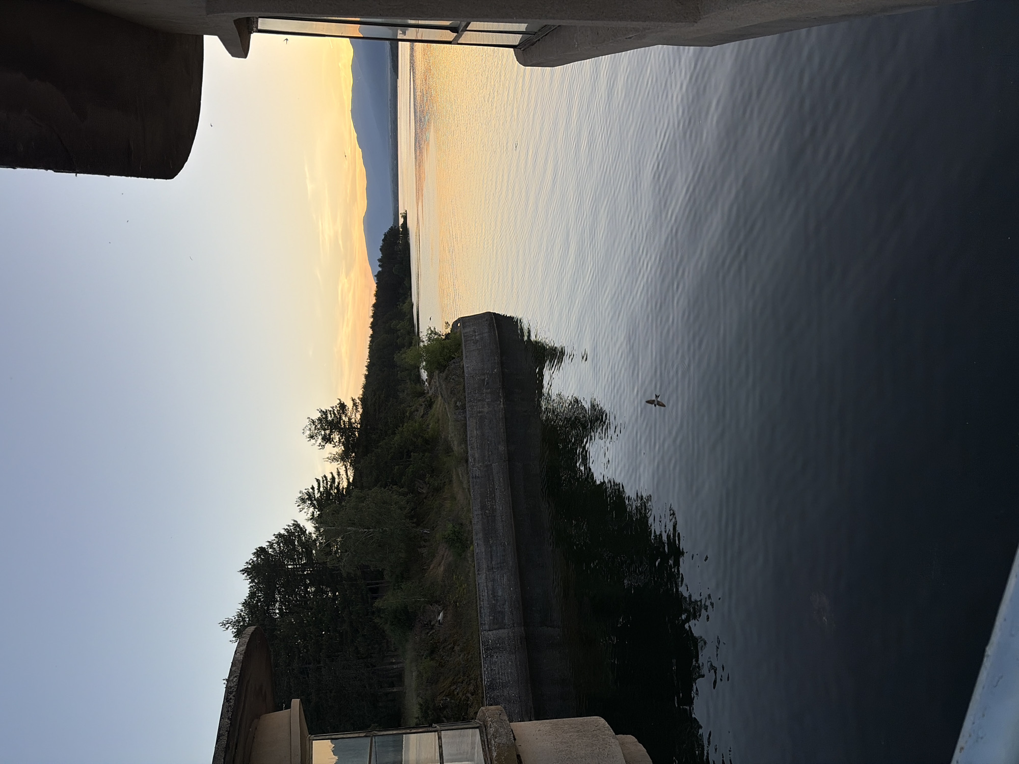The image depicts a scenic view of the Koprinka Dam in Koprinka, Bulgaria, at sunset. The foreground is dominated by a large body of dark water, with gentle ripples reflecting the fading light. To the left, a concrete structure, likely part of the dam's infrastructure, juts into the frame, featuring a window with a reflection of the sky. On the right, another concrete structure with a curved overhang partially obstructs the view, also with a window.
The middle ground reveals a concrete embankment along the water's edge, lined with trees. The trees are a mix of coniferous and deciduous species, densely packed on a sloped hillside. Beyond the trees, a vast expanse of water stretches towards the horizon, bathed in the warm glow of the setting sun. The sky transitions from a pale blue overhead to hues of orange and yellow near the horizon, where distant mountains are silhouetted against the vibrant sky. A single bird is visible in flight above the water.