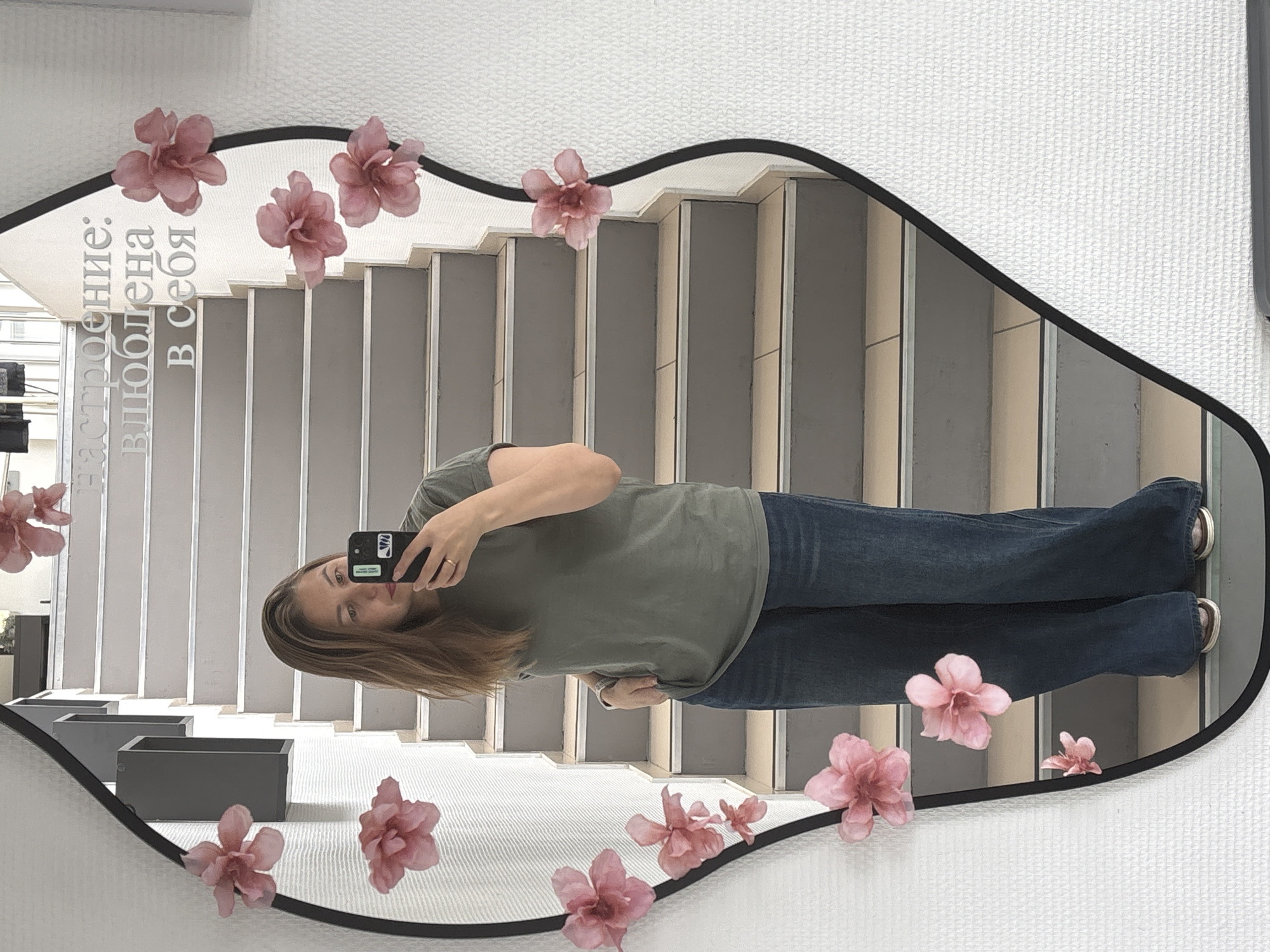 A mirror selfie of a woman with medium-length brown hair, wearing an olive-green T-shirt and blue jeans. She is holding a black smartphone with a white sticker on the back. The mirror, edged in black, is decorated with pink flowers. The reflection shows a staircase with gray steps, some gray planters, and what appears to be text in Russian near the top of the mirror reflecting: "настроение: влюблена в себя". The wall behind the mirror is textured white. The location is Chaikovsky, Russia.