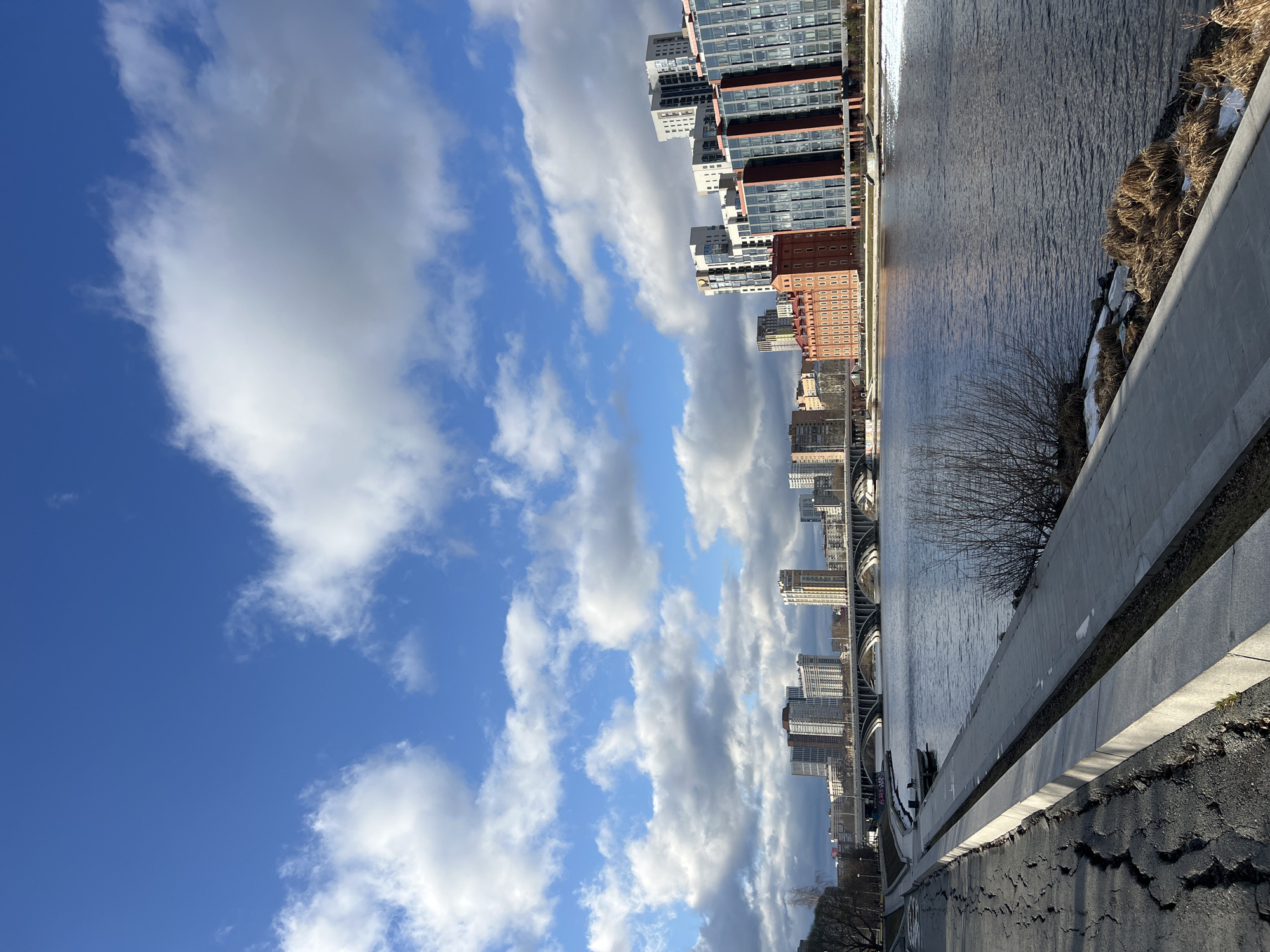 The image captures a view of the city of Yekaterinburg, Russia. A body of water runs parallel to a concrete embankment, beyond which lies the urban landscape. The cityscape consists of high-rise buildings, exhibiting various architectural styles. The sky is filled with a pattern of blue and white, showcasing a clear blue interspersed with scattered white clouds. The foreground presents a paved area with some visible cracks and wear. The overall composition suggests a bright, sunny day in an urban setting with a blend of natural and man-made elements.