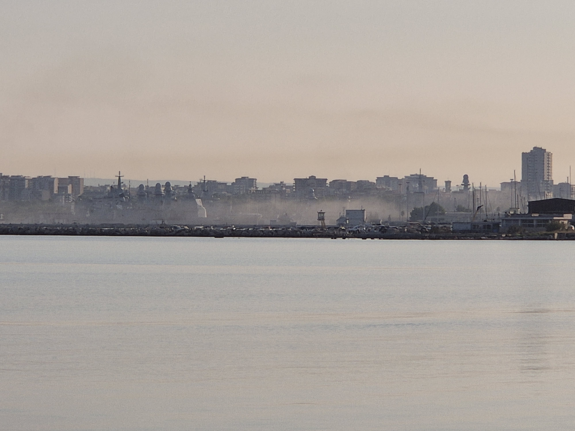 The image depicts a hazy, urban waterfront scene in Taranto, Italy. In the foreground, a body of calm water reflects the soft light of a hazy sky. A breakwater, constructed of large stones, separates the water from the city beyond. The middle ground reveals a dense collection of buildings and structures, characteristic of a harbor or industrial area. Numerous ships, including naval vessels and sailboats, are docked or anchored in the port. Tall buildings and urban structures rise behind the immediate port area, with a prominent, multi-story building visible on the far right. The overall atmosphere is subdued, likely due to atmospheric haze or mist, which softens the details of the cityscape.