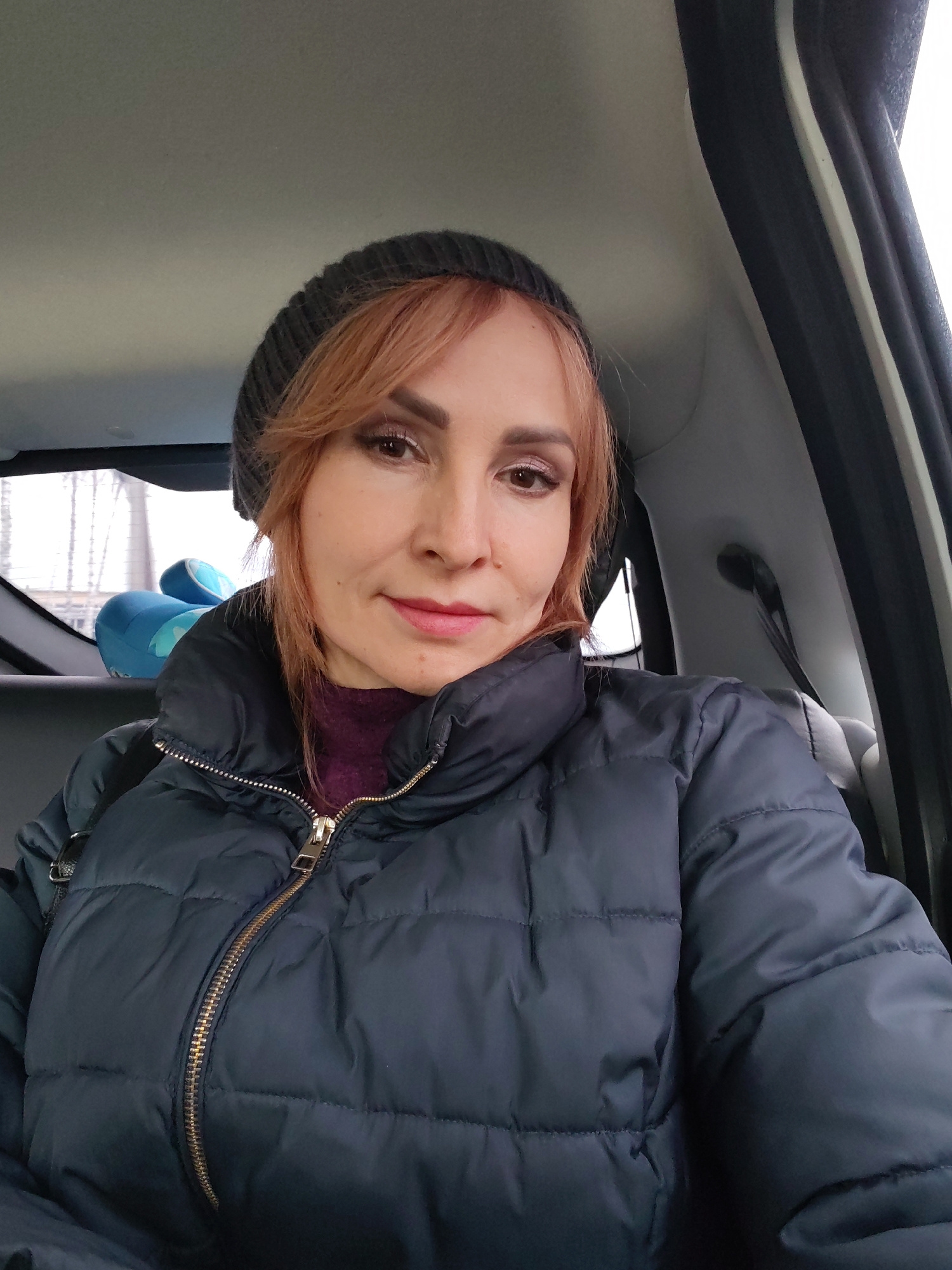 The image is a selfie of a fair-skinned woman with reddish-brown hair wearing a grey beanie and a dark blue puffer jacket. A maroon turtleneck sweater is visible under the jacket. She is likely inside a car, as suggested by the interior elements such as the door frame, headliner, and the back seat. Through the rear window, a blurred background reveals what appears to be a building or structure, possibly located in the city of Chaikovsky, Russia, along with what looks like a blue plush toy on the back seat. The lighting seems to be natural and diffused, indicating a daytime scene.