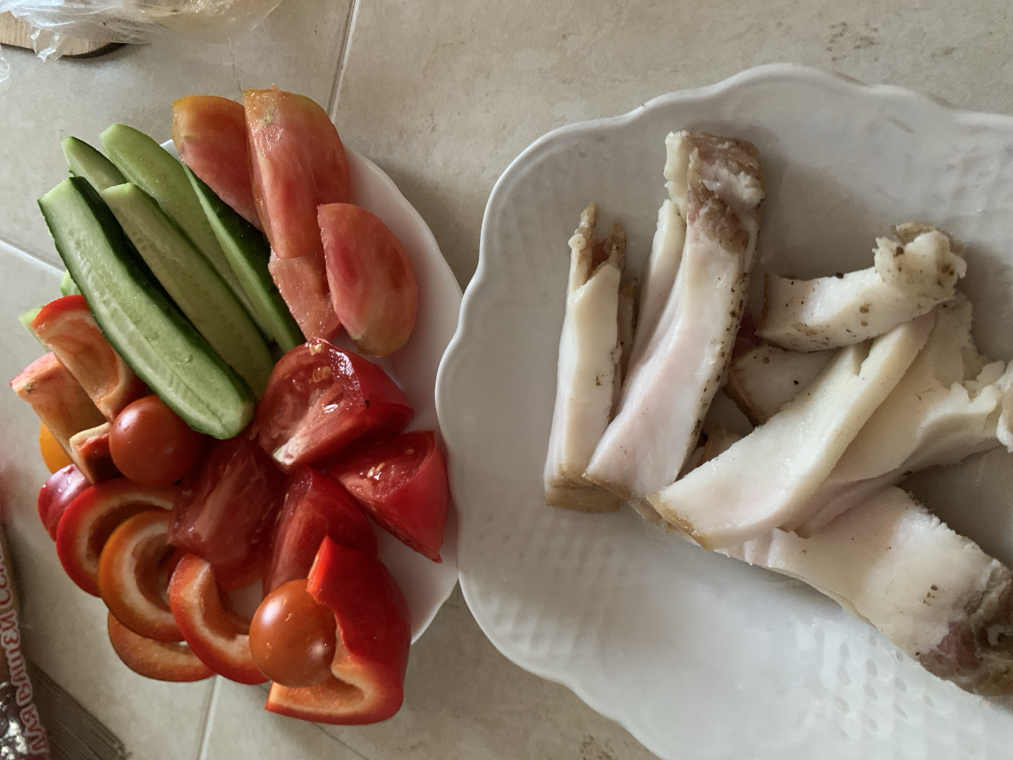 A high-angle medium shot shows two white plates of food on a light-colored tiled surface, possibly a kitchen counter, in Kapyĺ, Belarus. The upper plate contains a colorful assortment of sliced fresh vegetables, including red bell peppers, red tomatoes, and green cucumbers. The vegetables are arranged in a somewhat haphazard pile. The lower plate holds several slices of what appears to be cured pork fat, or salo, which is a traditional Belarusian food. The salo is white with streaks of pink and brown, and appears to be seasoned with black pepper. The lighting is natural, and the image is slightly out of focus in some areas.