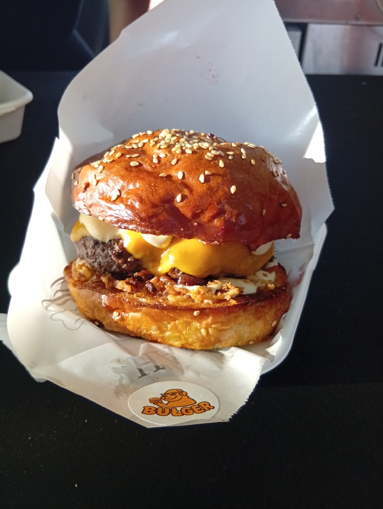 A close-up shot showcases a gourmet hamburger nestled in a white paper wrapper. The burger features a glossy, sesame seed-adorned bun, a thick beef patty, melted yellow cheese, and what appears to be crispy fried onions and sauce. A circular sticker with a cartoon bear wearing sunglasses and holding a burger, with the word "BULGER" visible, is affixed to the wrapper. The background is dark and out of focus, suggesting an outdoor or dimly lit environment. The scene is captured from a slightly elevated angle, with sunlight casting a shadow across the burger. The image's quality indicates it was taken with a mobile device. There is no discernible information within the image to determine its location in Wrocław, Poland.