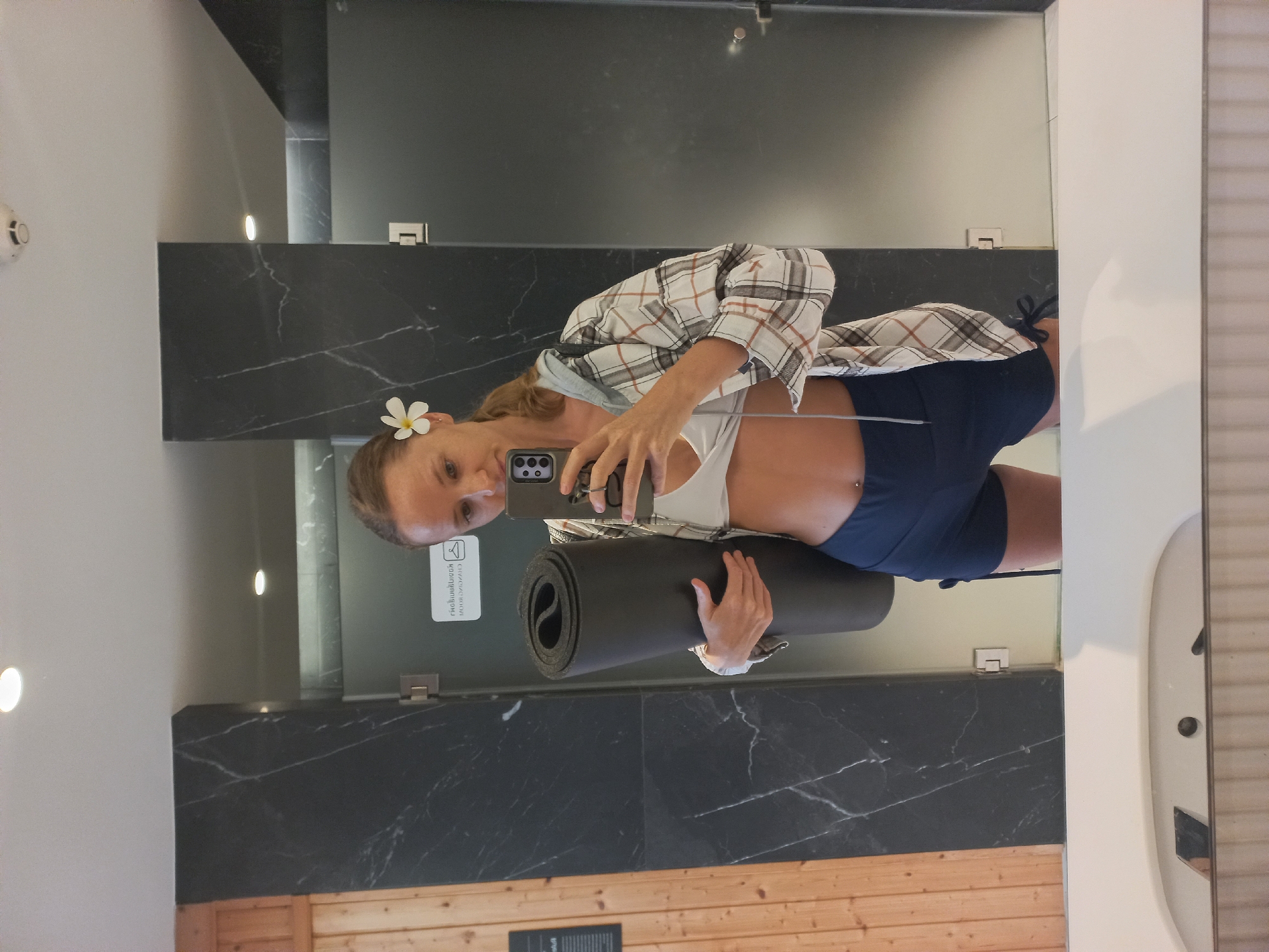 A woman is taking a selfie in a bathroom mirror. She has blonde hair, a flower in her hair, is wearing a sports bra and shorts, and is holding a rolled-up yoga mat. The bathroom has dark marble walls, a wooden floor, and a white wall. The selfie is taken in Pattaya City, Thailand.