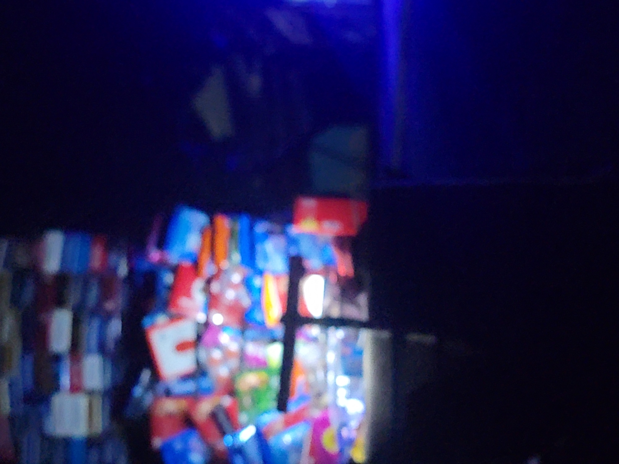The image depicts a poorly lit display of packaged goods, likely inside a store in Barden Yana, Nigeria. A dark, indeterminate background dominates the upper portion of the frame. The goods consist of colorful packages, possibly food items or household products, stacked or hanging on shelves. The items are somewhat blurry. A dark vertical and horizontal crossbar structure (possibly a window frame or shelf support) is visible in the middle ground.
