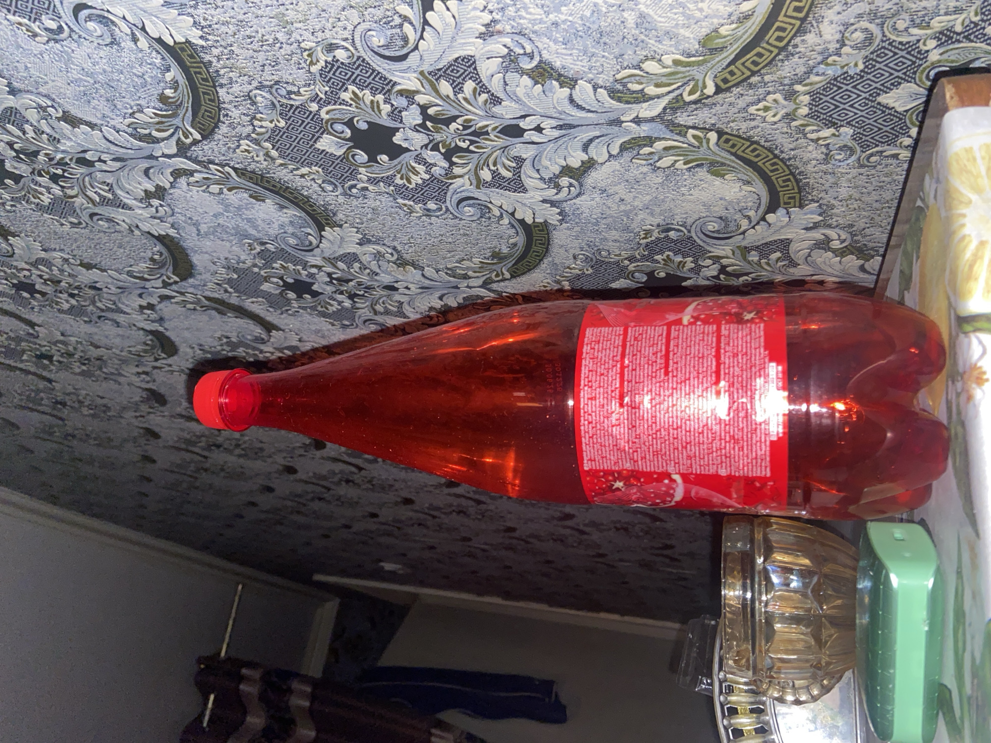 A red bottle of soda sits on a table in front of patterned wallpaper. To the left of the bottle, a small glass bowl and a green bar of soap are visible. The scene appears to be indoors, possibly in a residential setting.