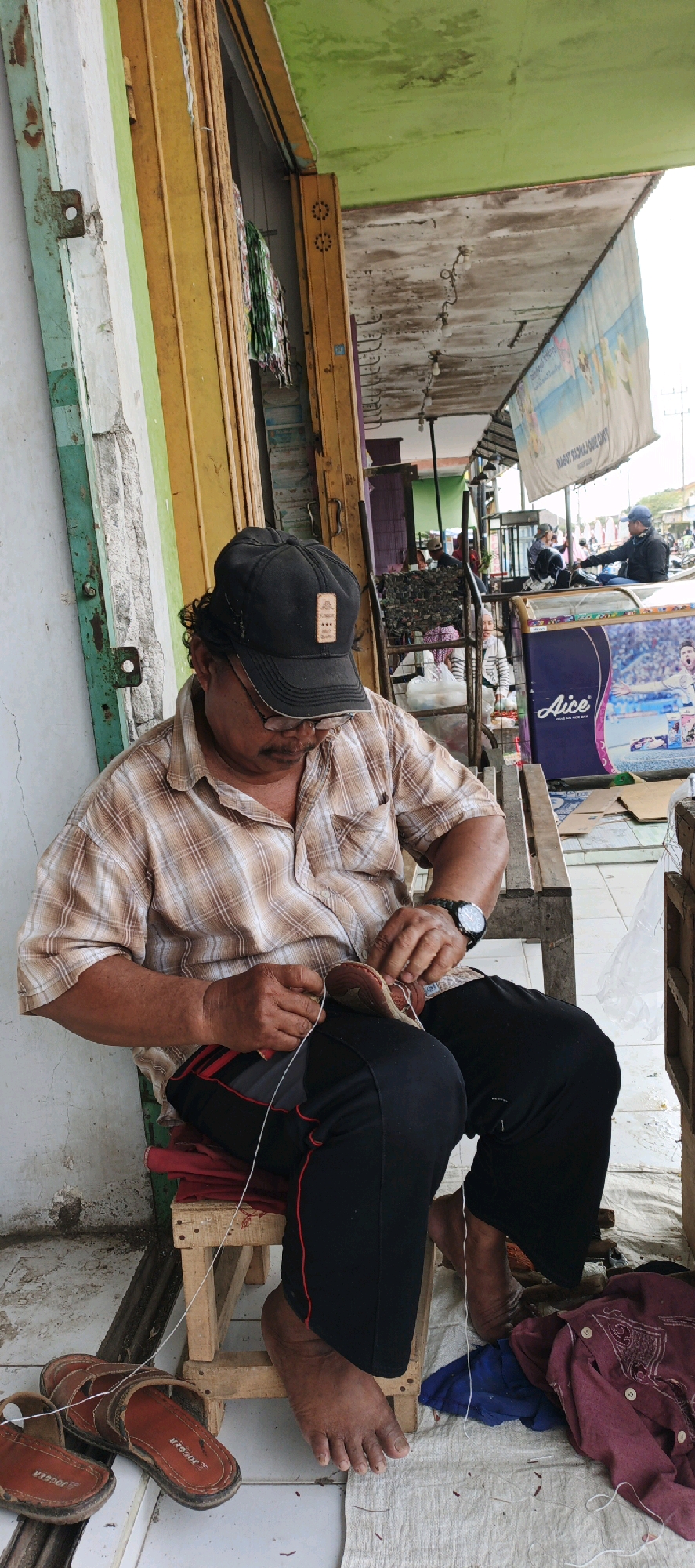 The image captures a scene in Kasiyan, Indonesia, where a man is seated outdoors, working on repairing a shoe. He is wearing a plaid shirt, black pants, and a black baseball cap, and has glasses on. The man is actively engaged in sewing with a needle and white thread, his focus directed downwards at the shoe he is holding. Beside him on the ground are a pair of brown leather sandals and a small red shoe. The setting appears to be a marketplace or a street-side stall, indicated by the presence of stalls with various goods and other people in the background, some of whom are also engaged in activities. The architecture suggests a local Indonesian environment, with corrugated metal roofing and wooden structures. The overall atmosphere is one of everyday commerce and craft.