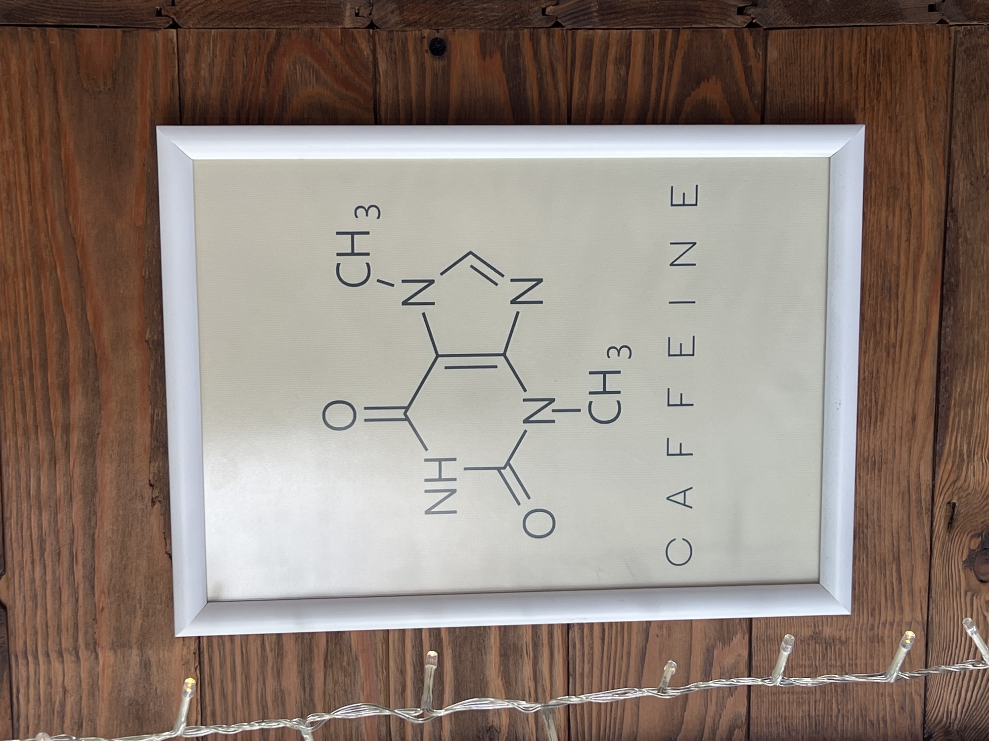 A framed picture depicting the chemical structure of caffeine along with the word "CAFFEINE" hangs on a brown wooden wall. The frame is white, and a string of small LED lights is visible below the frame. The location is unspecified.