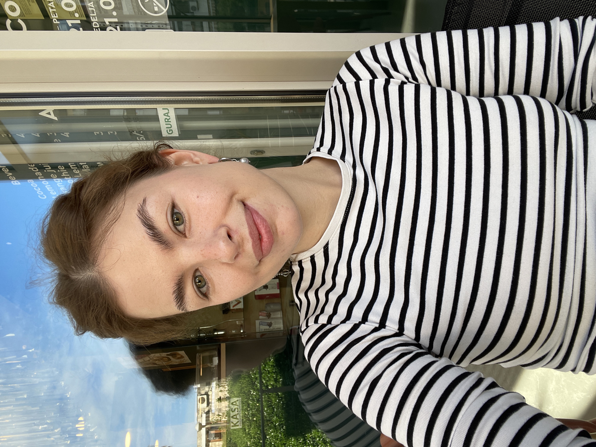 Here is a description of the image:
The photograph presents a young woman wearing a black and white striped long-sleeved shirt. She is facing the camera with a slight smile. The background is partially reflected in a glass surface, appearing to be a window or table, which shows an exterior scene, presumably of the Budva, Montenegro city surroundings. The reflection includes buildings with greenery and blue sky. The reflections from the glass surface show elements of the interior, including signage related to a cafe and some menu details that include menu items: Coca, lemonade, juice, smoothies, lemon, and other drinks. A sign displays the price 21.0. The words 'GURAJ' can also be read on a sign. The image is vertically oriented.