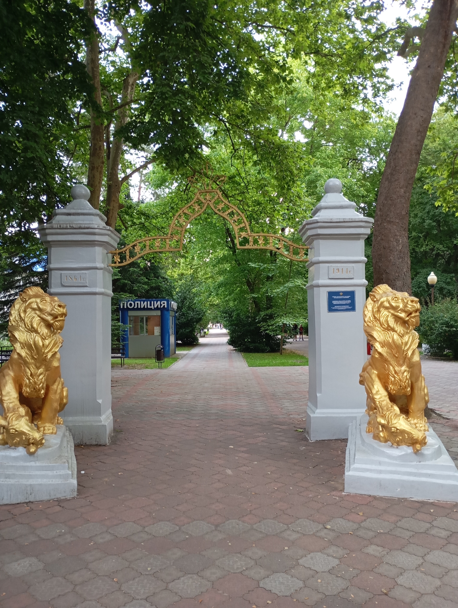 The image shows an entrance to a park in Goryachiy Klyuch, Russia, adorned with golden lion statues and an ornate archway. The pathway, paved with interlocking bricks, leads through a tree-lined avenue. On the left, a sign with the word "ПОЛИЦИЯ" (Police) indicates a small police booth. The date "1861г" is visible on the left pillar and "1911г" on the right pillar, likely historical markers. Lush green trees and foliage form a dense canopy overhead, dappling the scene with sunlight. The overall atmosphere is tranquil and inviting, suggesting a well-maintained public space.