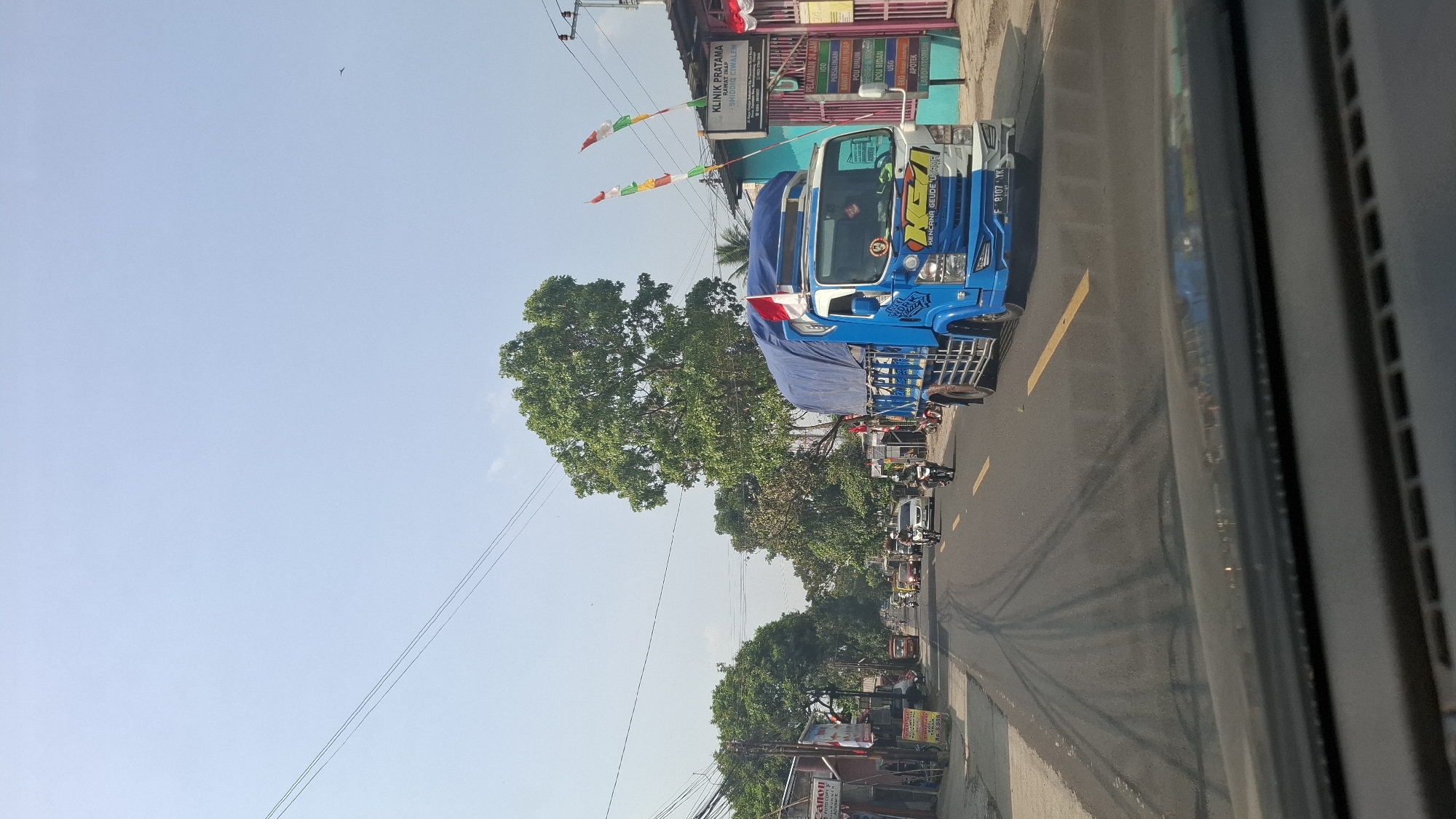 The image captures a street scene in Cikaroya, Indonesia, under a bright, clear sky. In the foreground, the dashboard of a vehicle is visible. The road ahead is paved and marked with yellow lines. A blue and white truck with a blue tarp covering its cargo is prominently positioned on the right side of the road, heading away from the viewer. The truck displays the Indonesian flag on its roof. Various other vehicles, including cars and motorcycles, are traveling on the road. On the left side of the road, there are buildings, including a clinic with signage that reads "KLINIK PRATAMA" and "SHIDDIQ CIVALEN". A large tree with lush green foliage stands to the left of the road, while another large tree is situated behind the clinic. Power lines are visible crisscrossing the sky. The overall atmosphere is one of a typical day in a bustling Indonesian town.