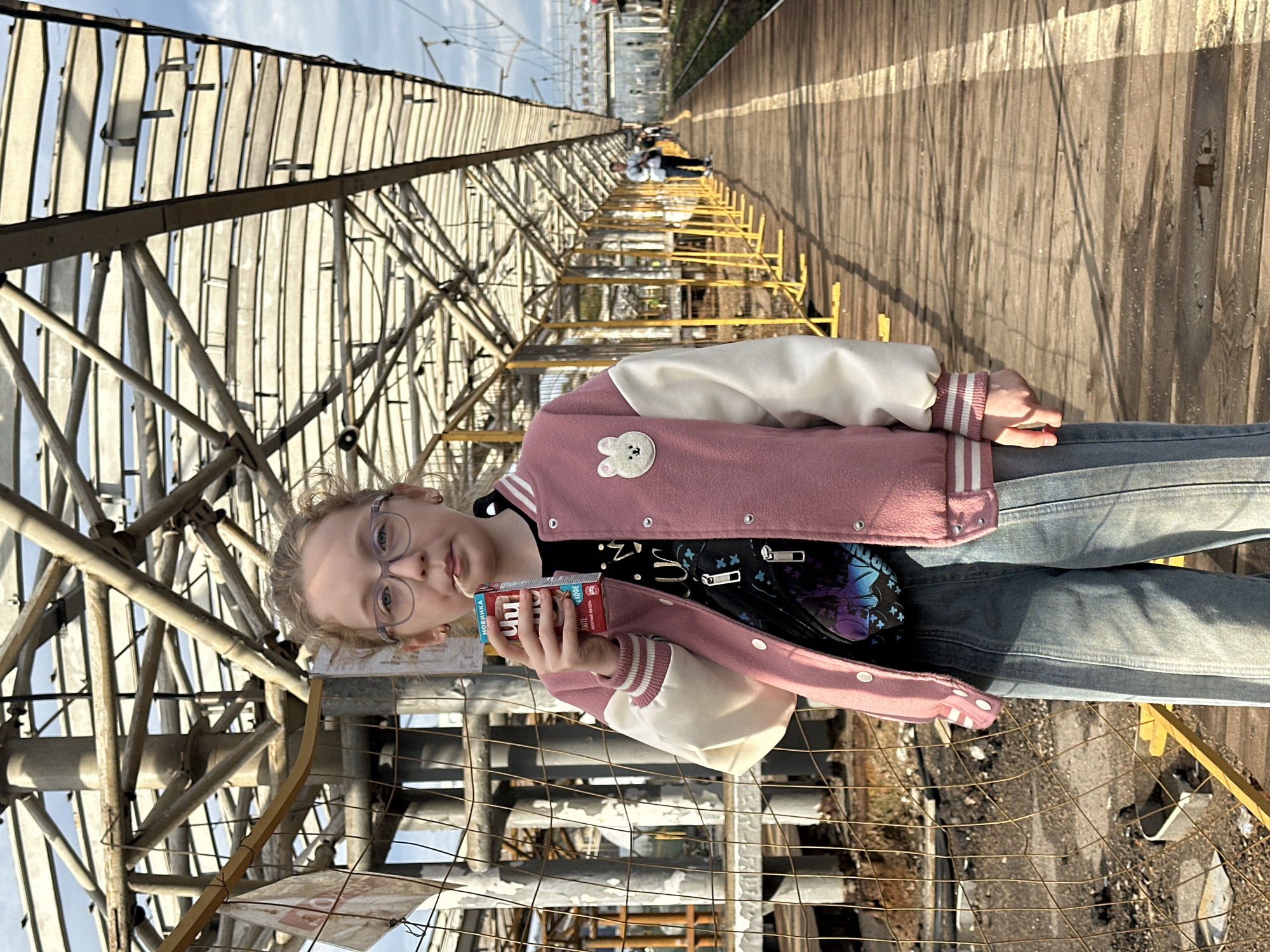 A young girl with blonde hair and glasses stands on a wooden platform, holding a juice box to her mouth. She is wearing a pink and white bomber jacket over a black t-shirt and light blue jeans. A small fanny pack with a cartoon design is worn across her chest. The platform appears to be a train station platform, with train tracks visible in the background. A covered walkway with a metal frame and translucent panels extends over the platform, and yellow safety barriers line the edge of the platform. In the distance, other people can be seen walking along the platform. The overall scene suggests a sunny day in an urban outdoor environment, likely a transportation hub. The presence of Russian text on the juice box and signs in the background indicate the location is Moscow, Russia.