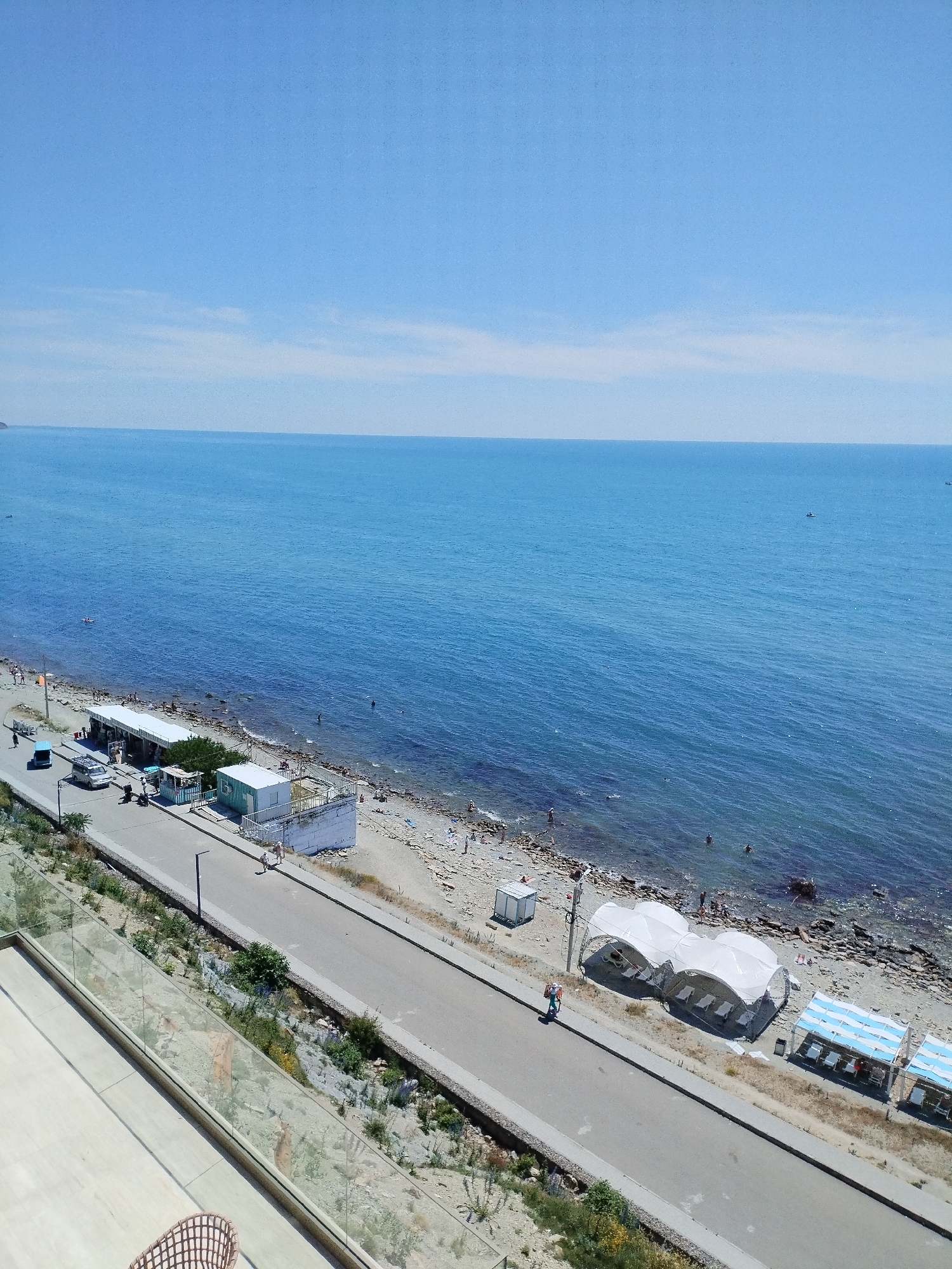 This is an aerial, wide-angle, full shot of a sunny day at a beach resort in Anapa, Russia. The scene captures the vast expanse of the blue Black Sea meeting a light blue sky. The shoreline is a mix of sand and pebbles, with a road running parallel to it.
Along the beach, several white, tent-like structures are set up, likely for shade. Some beach chairs are visible beneath these structures. There are a few people scattered on the beach, some swimming in the sea and others sunbathing. A small boat is visible in the distance on the water.
The road has a few cars and motorcycles parked along its side, as well as some small buildings that appear to be kiosks or changing rooms. A few individuals are walking or cycling along the road and the beach path. The foreground shows a modern balcony with a glass railing, overlooking the beach and sea, suggesting the vantage point is from a hotel or residential building. The vegetation is sparse, with some dry grasses and low-lying plants along the embankment. The overall atmosphere is one of a typical summer day at a seaside resort.