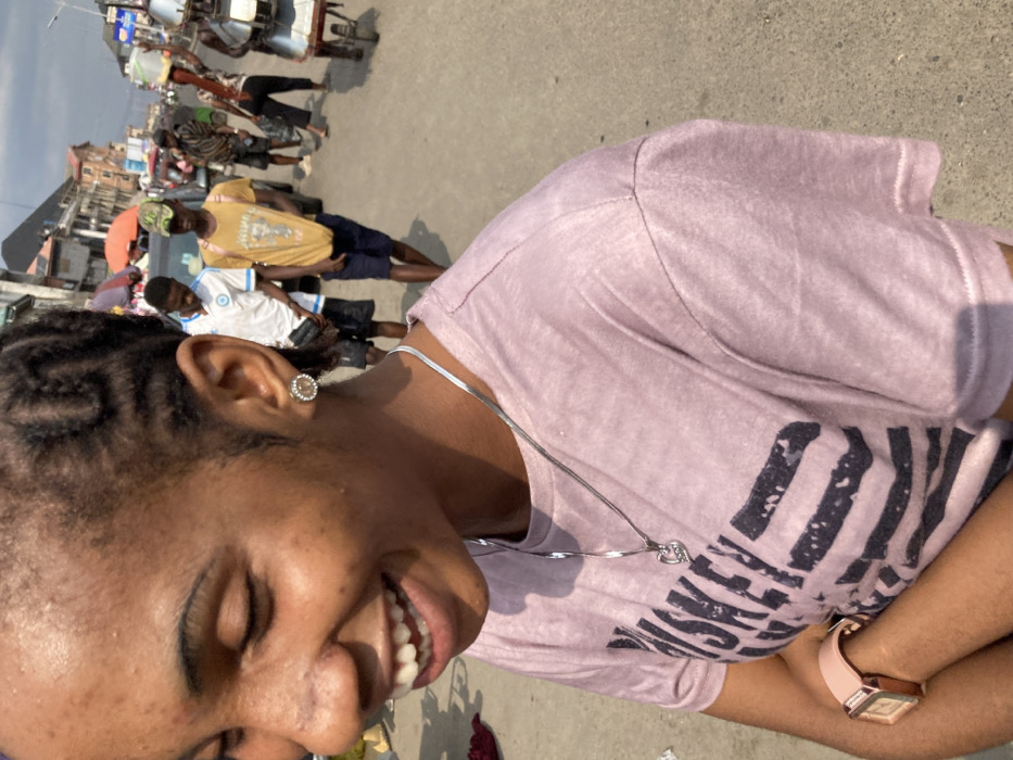 The image depicts a female individual in the foreground, smiling broadly with closed eyes, revealing her upper and lower teeth. She has dark skin, braided cornrow hair styled in a pattern, and wears a circular stud earring in her visible right ear. A silver-colored flat chain necklace with a heart-shaped pendant is around her neck. She is wearing a light purple t-shirt with a faded dark graphic print, partially visible as "WHISKEY" and a striped design. A light pink square-faced wristwatch is visible on her left wrist, and her arms appear crossed.
The scene is an outdoor urban street or market environment in Ura Nta Umuarandu, Nigeria. The ground surface is paved. In the midground and background, multiple individuals are present, predominantly male, engaged in walking or standing. Notable figures include a male in a white t-shirt with blue trim, another in a yellow t-shirt and camouflage cap carrying a backpack, and a person further back carrying a large metal container on their head. A wheeled cart is also visible on the far right. Multi-story buildings with various roof types are visible in the distant background, and elements suggesting market activity, such as canopies or stalls, are present. The scene is illuminated by bright daylight.