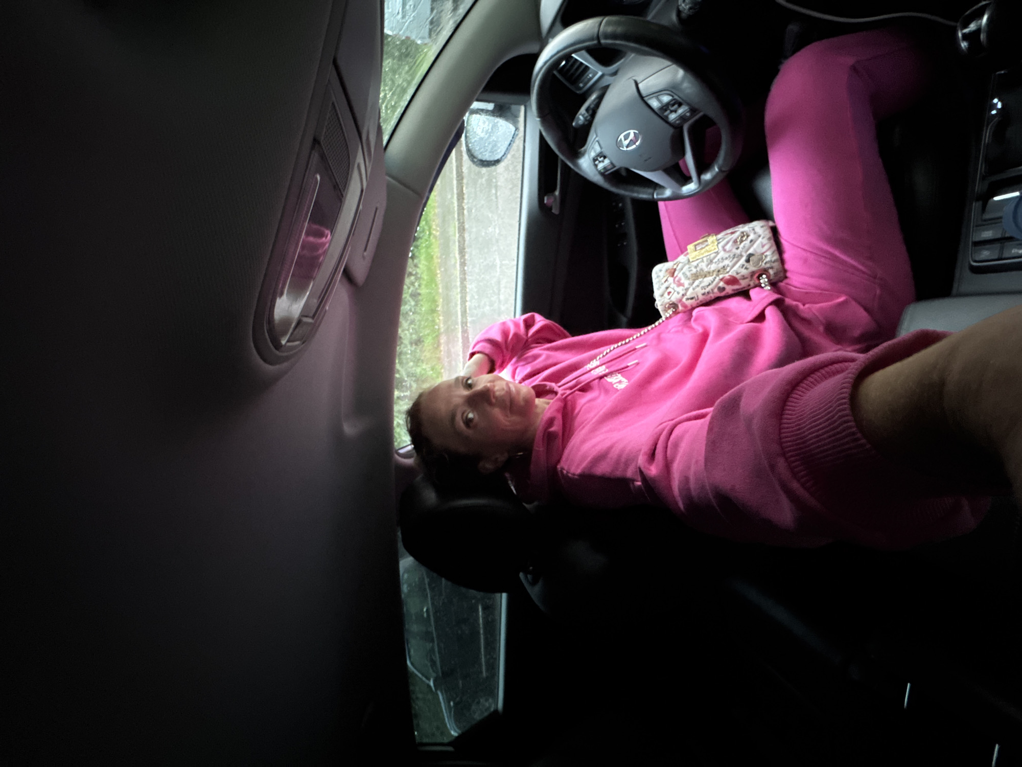 An overhead wide shot shows a woman in a bright pink tracksuit sitting in the driver's seat of a car. The woman is facing the camera, her right hand resting on her head. Her left arm is extended towards the camera. A small, patterned purse with a gold chain strap is resting on her lap. The car's interior is visible, including the steering wheel and dashboard. Outside the car, it appears to be raining, with raindrops visible on the windshield and side windows. In the background, another dark-colored vehicle is visible parked on a street. The image appears to be a selfie, taken from the perspective of the person in the driver's seat.