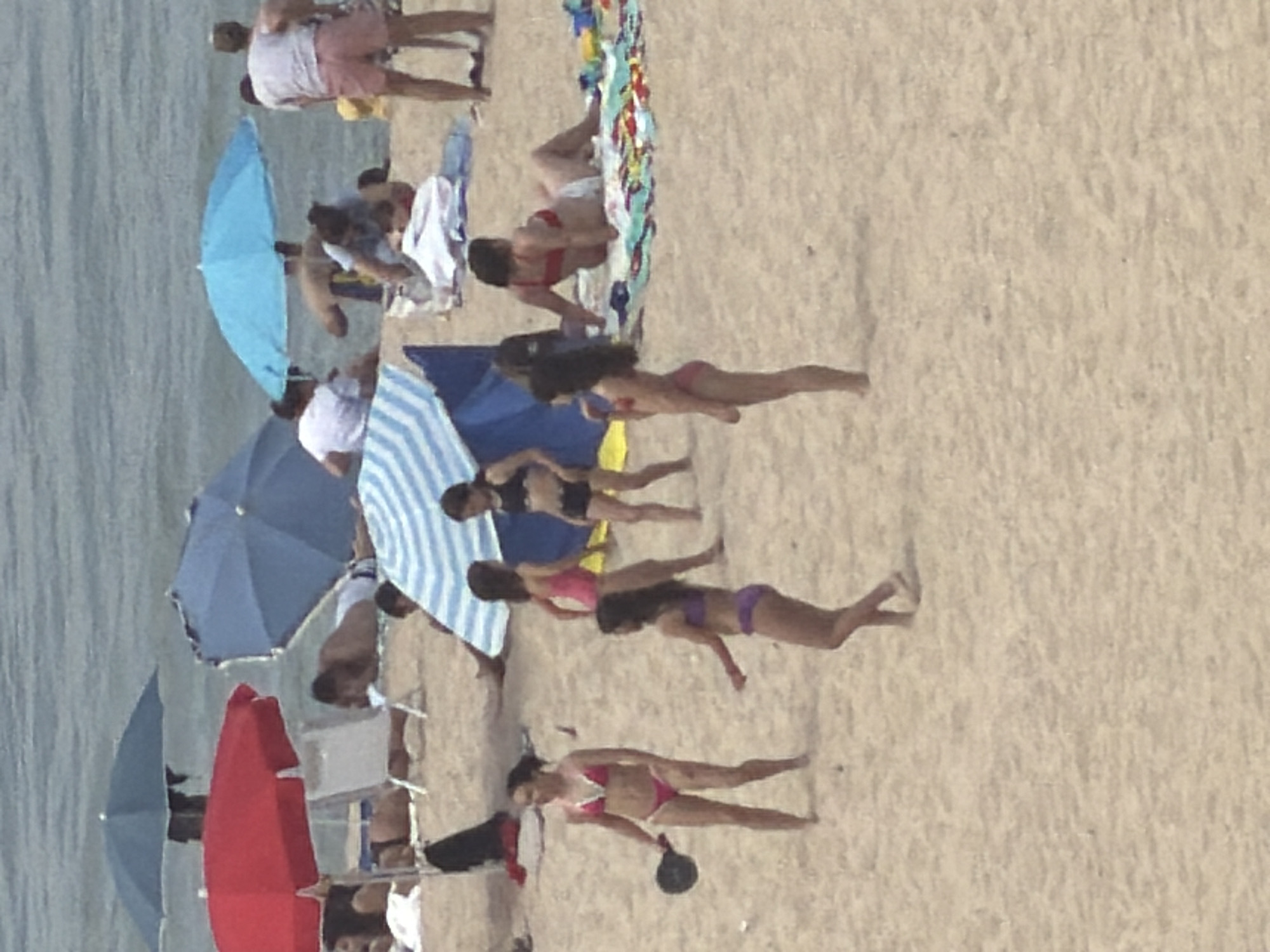 The image depicts a crowded beach scene on a sandy shore with the ocean in the background. Several people are visible in swimwear, engaged in various activities. In the foreground, a woman in a pink bikini stands holding a black object, possibly a small ball or paddle. To her right, another woman in a purple bikini is visible. Further back, a woman in a pink bikini stands with her back to the camera, and to her right, a woman in a black bikini is also visible. Scattered throughout the beach are several beach umbrellas in red and blue. Some individuals are lying on towels or chairs, while others are standing or walking. The overall atmosphere suggests a sunny day at the beach with many visitors. The location is identified as the city of Espinho, Portugal.