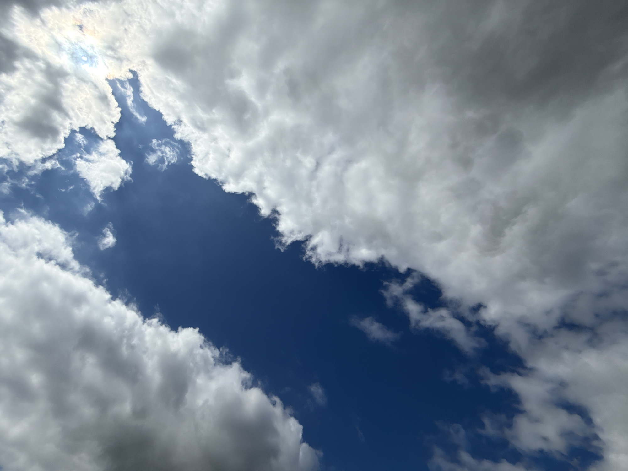 The image is a close-up, low-angle shot of a sky filled with clouds. A large gap in the clouds reveals a deep blue sky. On the right side, the sun is visible, peeking through the clouds, causing a slight lens flare with some iridescent colors. The clouds vary in texture and color, with some appearing bright white and fluffy, while others are darker and more ominous. The overall impression is of a dynamic sky, possibly on a partly cloudy day. The image does not contain any recognizable landmarks, people, or specific objects that would indicate a location within Großpösna, Germany, or any other specific city.