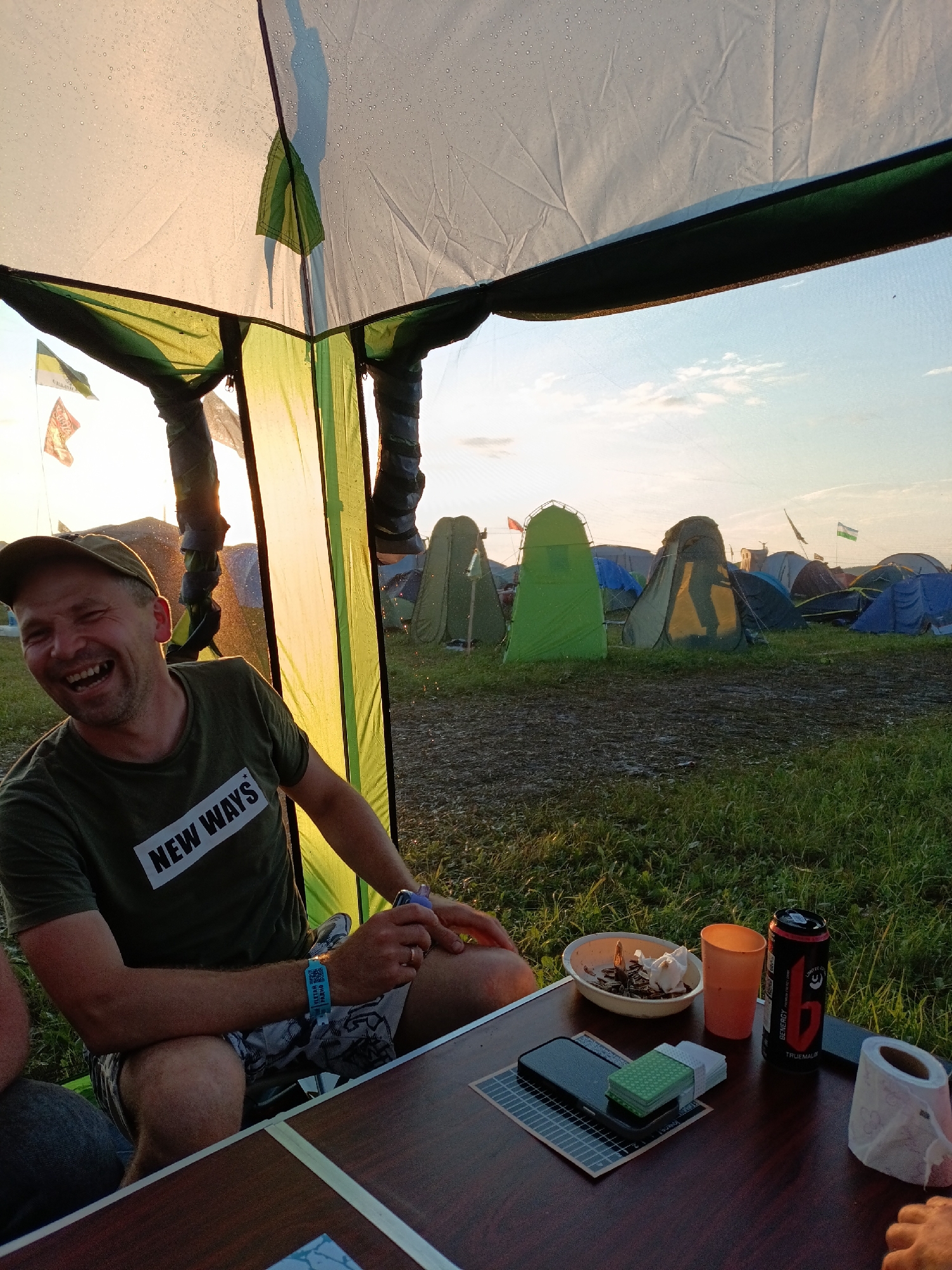 The image depicts a man wearing a green t-shirt with "NEW WAYS" printed on it and a baseball cap, laughing while seated at a table in what appears to be a campsite. The campsite is filled with numerous tents, and the ground is covered in grass and some muddy patches. The sky is visible in the background, with the sun setting, casting a warm glow. On the table, there's a bowl containing food, an orange cup, a can of "BENERGY" energy drink, a mobile phone, a packet of tissues, and a roll of toilet paper. The man has a festival wristband on his left wrist. This scene is set in a rural or outdoor festival environment, not specifically in the city of Lagunovo, Russia.