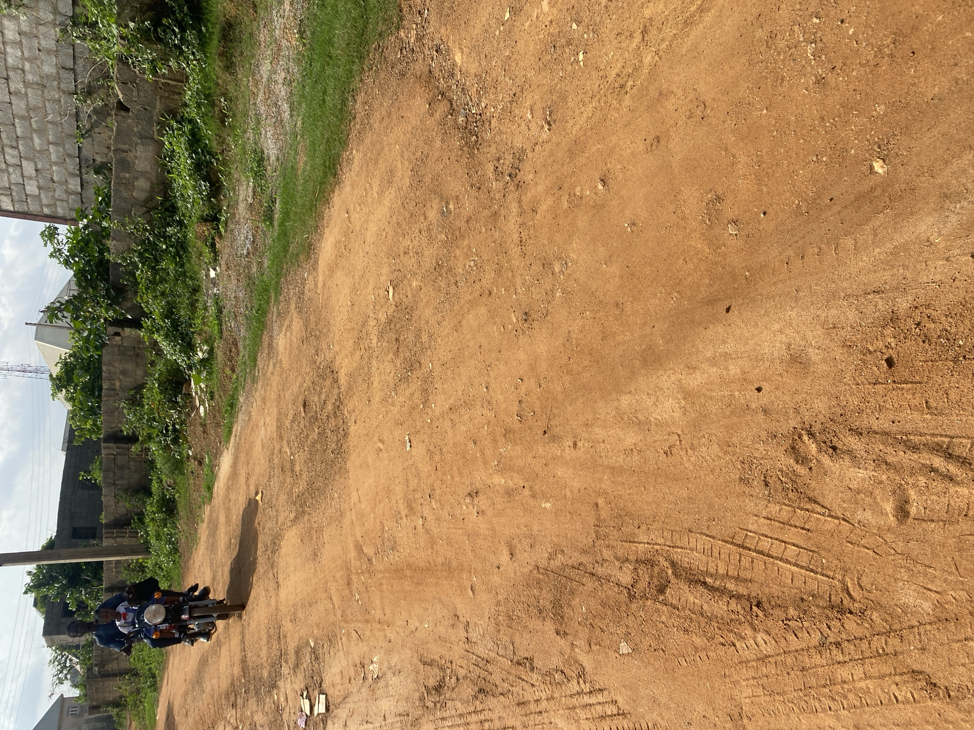 An adult male is riding a motorcycle on a dirt road in Dam Camp, Nigeria. He is wearing a dark blue shirt and pants. A young child, wearing a white shirt and a red bowtie, is seated in front of him on the motorcycle. They are both looking forward. The road is made of orange dirt and has tire tracks. There is green grass and vegetation on the right side of the road. In the background, there are buildings and a wall. The sky is cloudy.