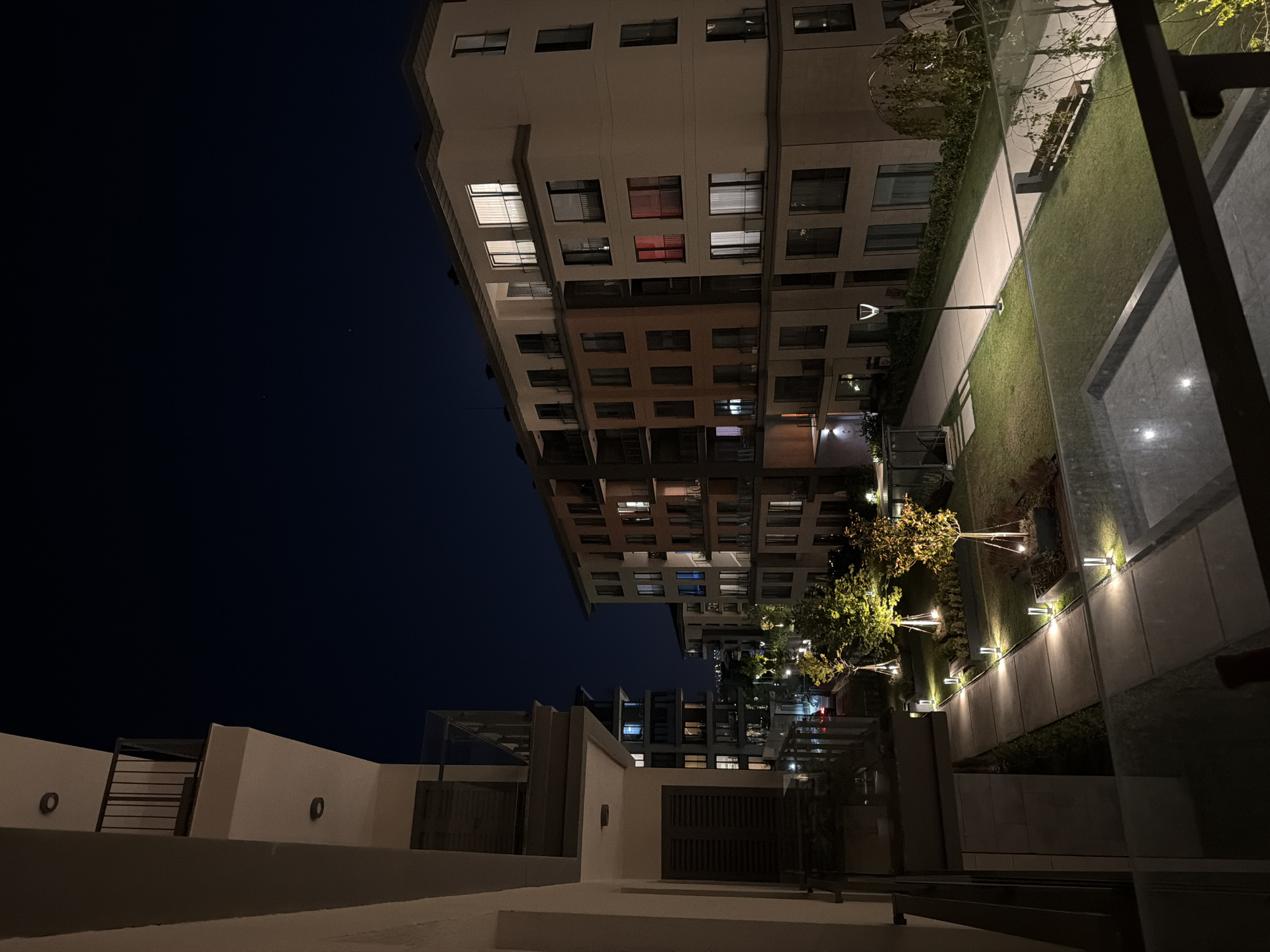 This is an outdoor, nighttime photograph taken from a balcony in the Çekmeköy district of Istanbul, Turkey. The view looks out onto a courtyard area between apartment buildings. In the foreground, a glass balcony railing is visible. To the left, a portion of a light-colored apartment building with balconies and shutters is shown. In the center and to the right, a modern apartment complex is depicted with multiple stories, balconies, and numerous windows, some of which are illuminated, casting light into the night. One window on the right-hand building has a distinctive red glow.
The courtyard features a paved walkway with integrated lighting, flanked by manicured lawns. Several young trees are planted in raised beds and illuminated by spotlights, creating a warm glow on their foliage. A park bench is situated on the lawn in the middle ground. The sky above is dark blue and clear, with a few faint stars visible. The overall impression is one of a quiet, residential urban environment at night. There are no people visible in the frame.
