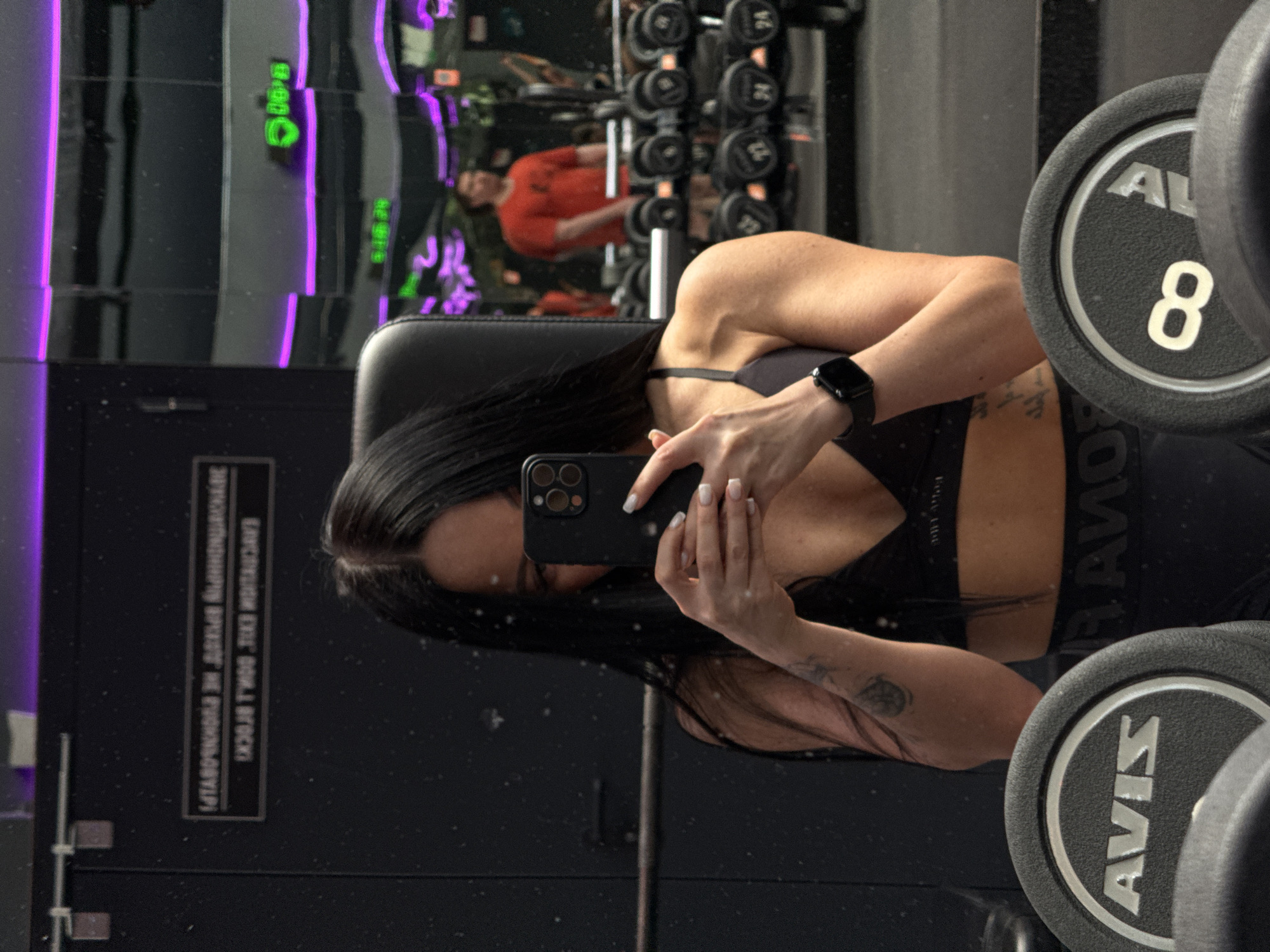 An image captures a woman taking a mirror selfie in a gym, with the camera held to obscure her face. She has long, dark hair, wears a black sports bra and athletic bottoms, and has a smartwatch on her left wrist. A tattoo is visible on her right arm. The reflection in the mirror shows a gym interior with various equipment.
In the background reflection, a man in an orange shirt is visible. Numerous dumbbells are racked, and a prominent dumbbell in the foreground clearly shows the number "8" and the brand "AVIS". The gym features sleek, dark surfaces and atmospheric purple and green lighting elements, possibly neon or LED strips.
On a dark panel or door reflected in the mirror, Cyrillic text is visible. When read correctly (as if not reflected), it states: "ЭКСПЛУАТАЦИЯ ЗАЛА С 9.00 ДО 22.00 БЕЗ ПЕРЕРЫВОВ И ВЫХОДНЫХ," which translates to "Gym operation from 9:00 to 22:00 without breaks and weekends." The setting is an indoor fitness facility, likely located in Kazan, Russia.
