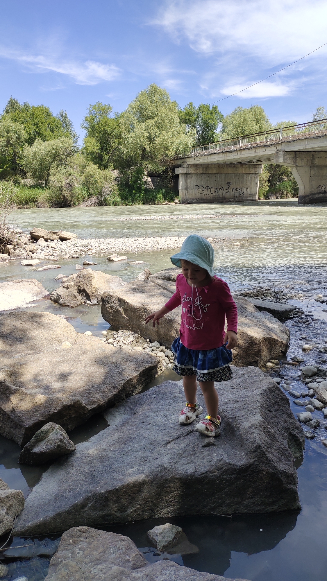 In Taldykorgan, Kazakhstan, a young girl wearing a pink long-sleeved shirt, a blue skirt, and a light blue sun hat stands on large rocks by a river. The river is shallow with a rocky bed, and water flows gently. Behind the river, a concrete bridge spans the water, with trees and greenery on the riverbanks. The sky is partly cloudy. Graffiti is visible on the underside of the bridge. The girl appears to be interacting with the environment, with one hand extended towards the water.