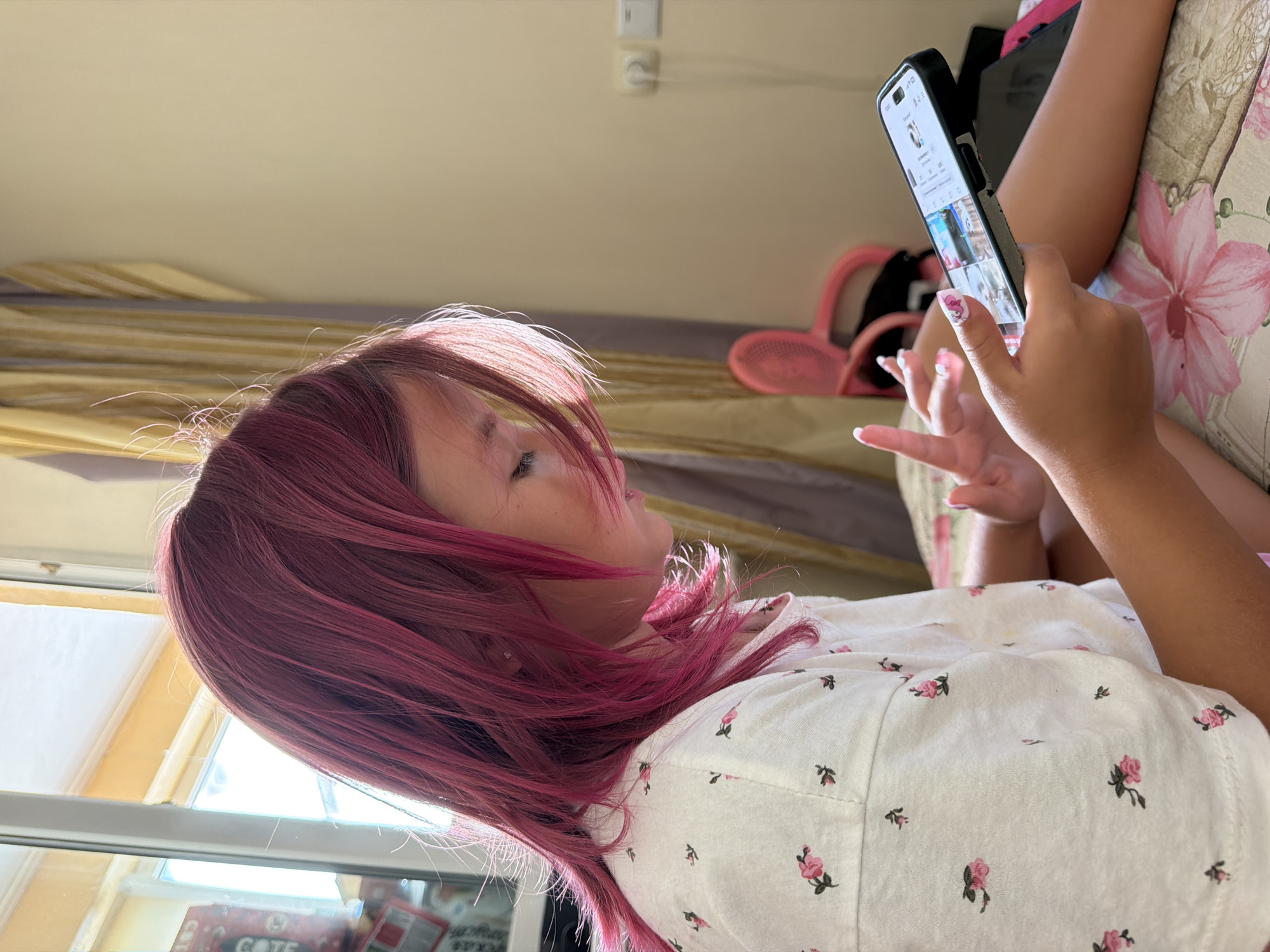A young girl with bright pink hair, wearing a white t-shirt with a floral pattern, is seated on a bed and looking at a smartphone. The room appears to be a bedroom, with a window that lets in natural light. Curtains are visible in the background, as is a pink plastic chair. The phone screen displays a social media feed. There is no indication of the city of Partenit, Ukraine, in this image.