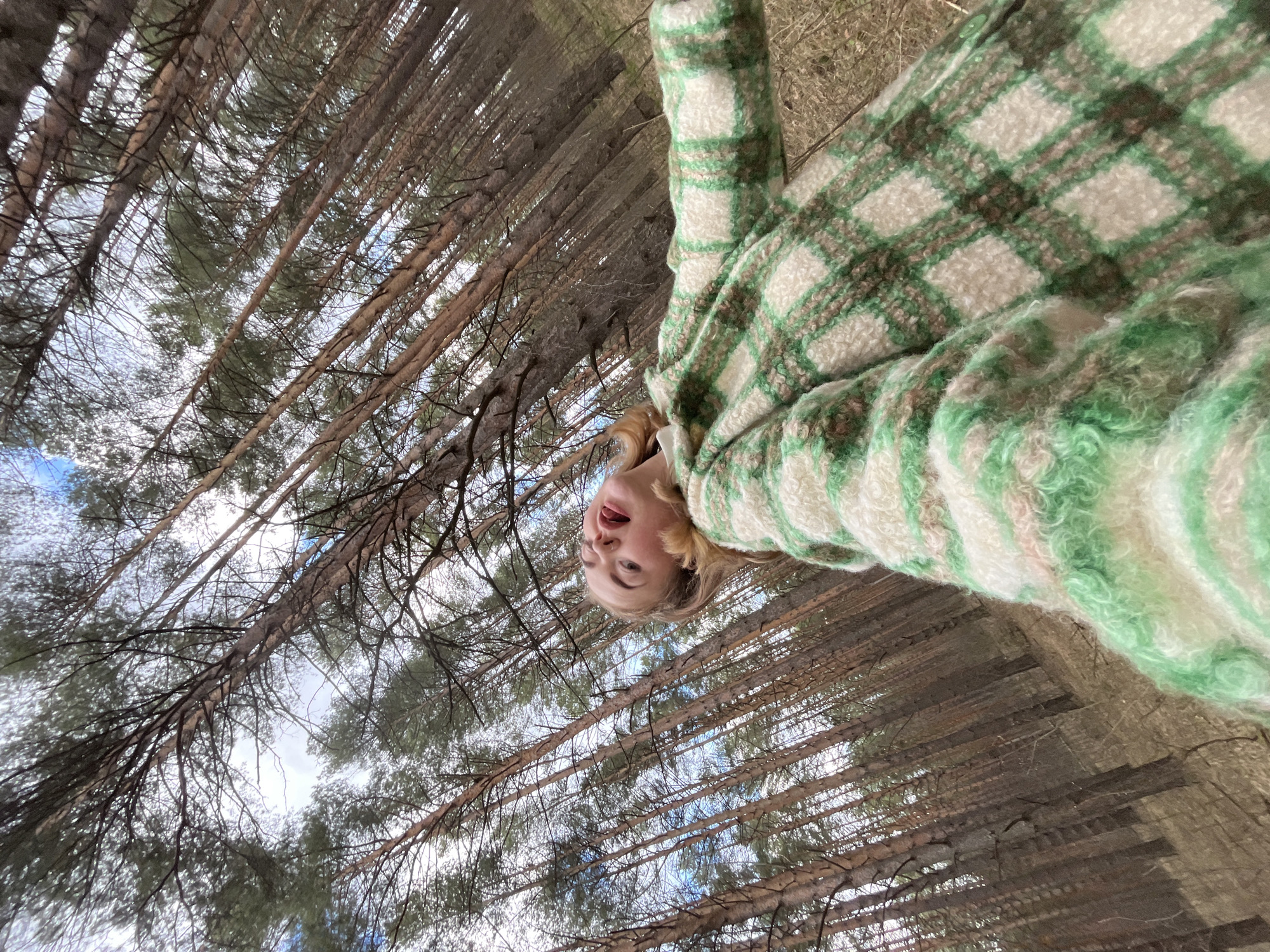 A young person is pictured from a low angle, looking upwards towards the camera with a playful expression. Their mouth is open, revealing teeth and tongue, suggesting a moment of surprise or amusement. They have light blonde hair and are wearing a thick, fuzzy jacket or coat. The jacket features a prominent plaid pattern in alternating squares of light green, darker green, and off-white/cream.
The setting is a dense pine forest, with numerous tall, slender tree trunks and branches extending vertically upwards around the subject. The camera's upward perspective captures the intricate network of branches against a bright sky, which appears partially cloudy in some areas. Sunlight filters through the tree canopy, creating a natural, bright daytime scene. The image conveys a cheerful and immersive outdoor experience within the forest. No text is visible in the image. The location context of Chaikovsky, Russia, aligns with the appearance of a pine forest environment.