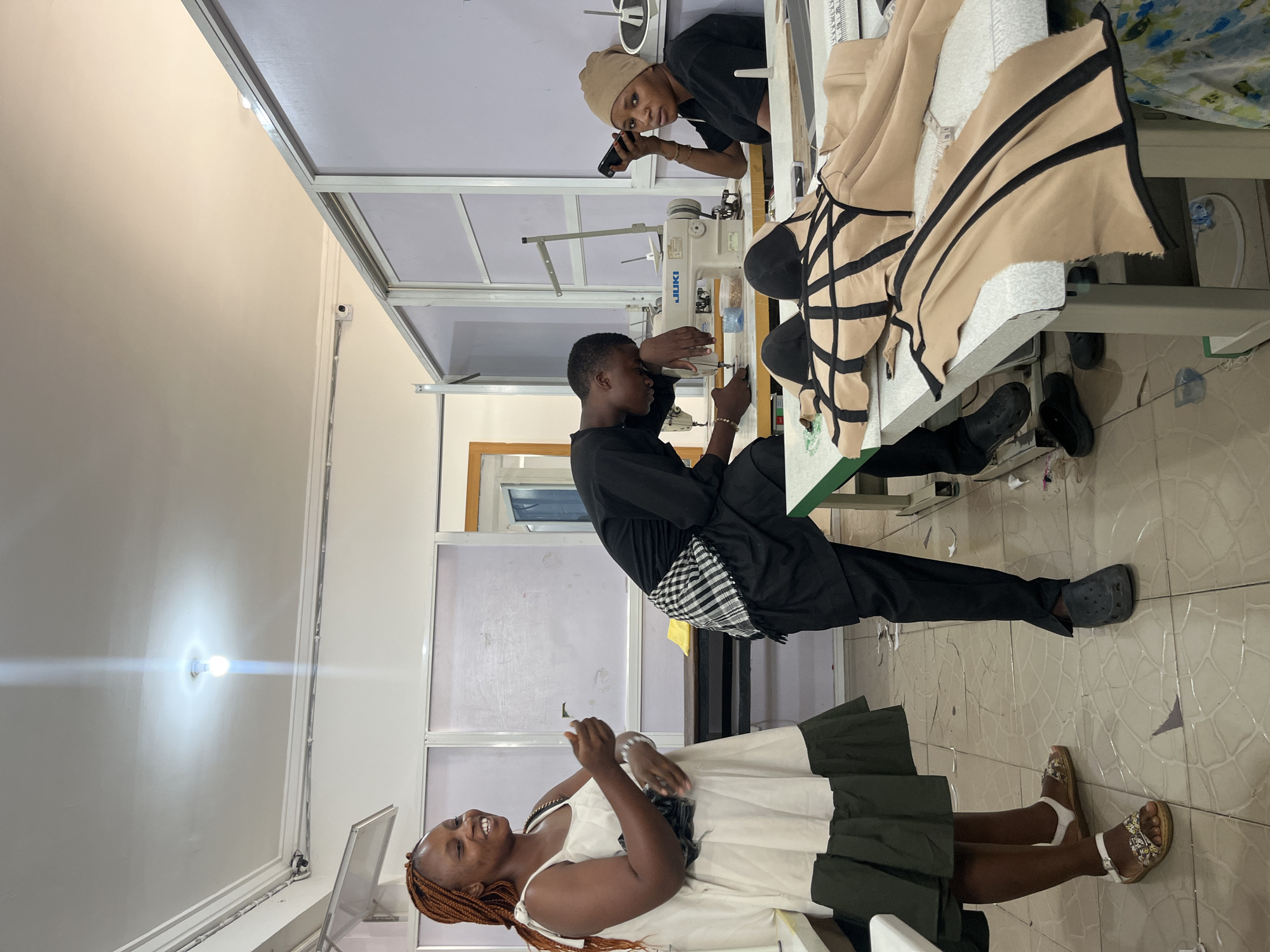 This indoor portrait image, captured from a slightly elevated perspective, depicts three individuals in what appears to be a fashion design workshop or tailoring studio.
In the upper part of the frame, a woman wearing a black t-shirt and a beige head covering looks directly towards the camera, holding a black smartphone in her right hand. She is positioned behind a sewing machine table, observing the activities.
In the middle, a young man, dressed in a black long-sleeved top and black trousers with a black and white checkered fabric tied around his waist, is actively working at a sewing machine. The brand "JUKI" is visible on the machine. He is focused on manipulating fabric under the needle, with a piece of beige fabric featuring bold black stripes draped over the machine table.
In the foreground and lower part of the image, another woman with braided hair, wearing a white sleeveless dress with a dark green pleated skirt section, smiles broadly while looking slightly upwards and to her left. Her hands are raised slightly, suggesting she might be talking or clapping.
The setting features white walls, metal-framed glass partitions, and a tiled floor. A bright, wall-mounted light fixture casts a strong glare in the left background. Various sewing tools and fabric scraps are subtly visible around the workstation. One pair of black Crocs and a pair of white and brown sandals are visible on the floor near the individuals.
The overall atmosphere suggests a collaborative or instructional environment, possibly a training session or a busy workday in a creative space. The time of day cannot be determined, as the lighting is predominantly artificial.