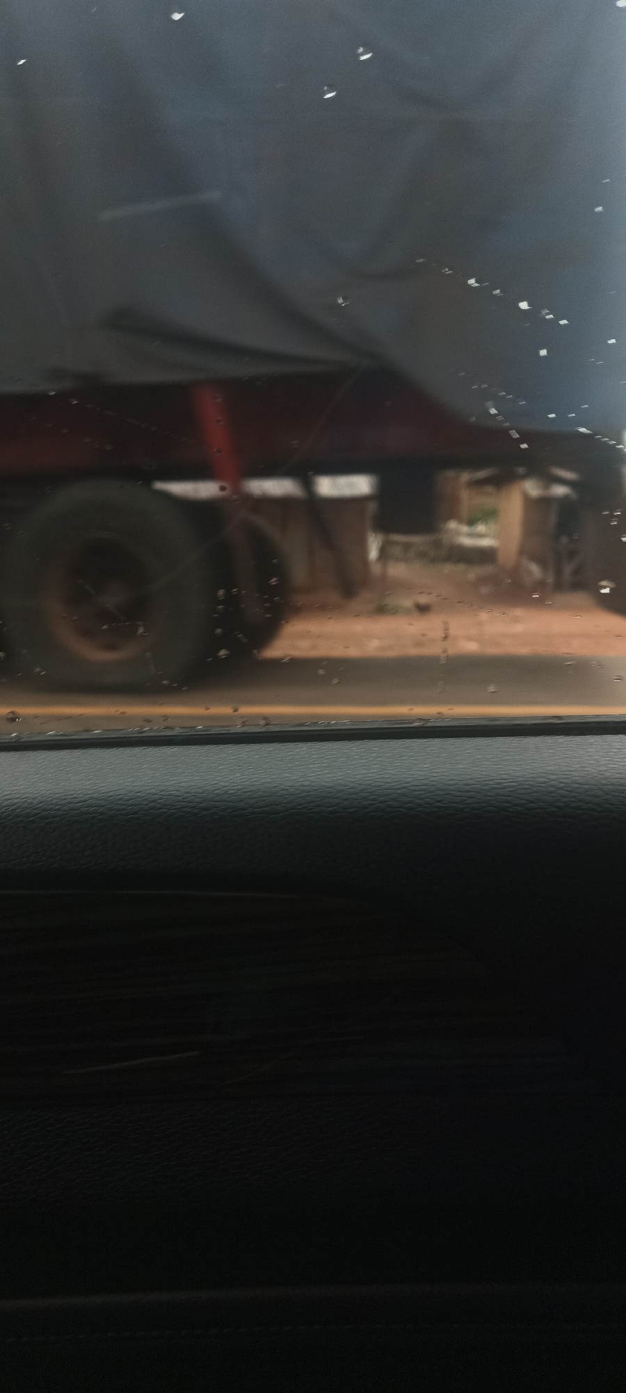 The image shows a view from inside a vehicle, looking out through a rain-splattered window. A large truck is partially visible, with its red chassis and large tire prominent in the frame. Behind the truck, a glimpse of a building or structure can be seen. The environment appears to be outdoors, with a dirt or gravel road visible. The dashboard of the vehicle occupies the bottom portion of the image, suggesting the photo was taken from the passenger seat. I do not see any evidence of Gwadanye, Nigeria. I do not see any people.