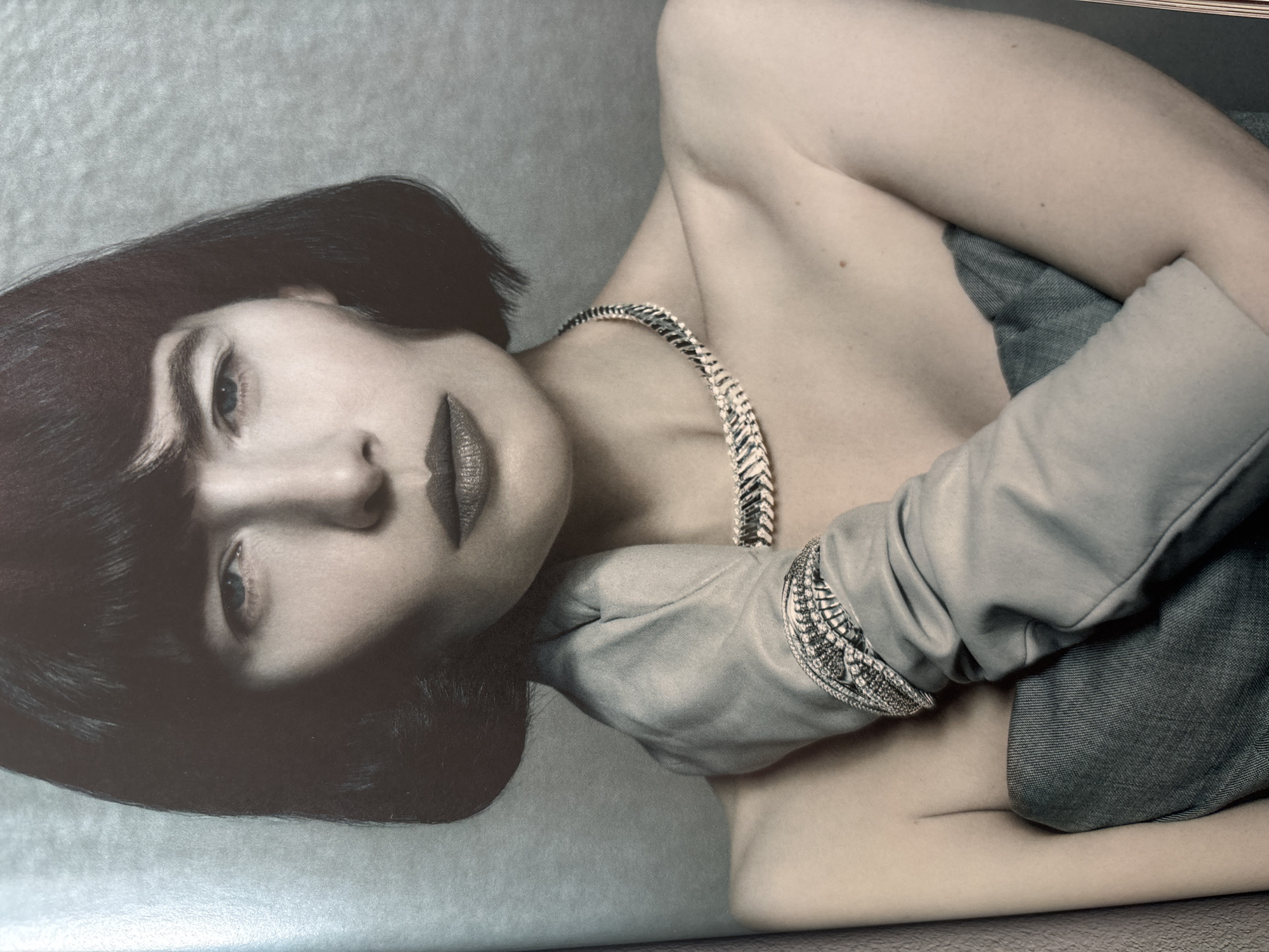 The image is a black-and-white portrait of a woman with short, dark hair and full lips. She is wearing a necklace and a light-colored garment that appears to be a glove or sleeve. Her arm is raised and crossed over her chest. The background is a neutral, light gray color. The image does not contain any information to identify a location in the city of Vsevolozhsk, Russia.