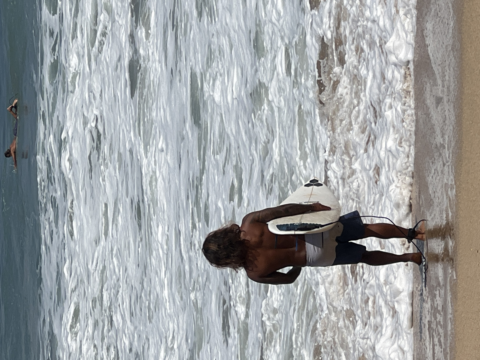 A surfer with long, dark hair and tattoos on his arm stands facing the ocean, holding a white surfboard. He wears grey shorts and is barefoot. In the distance, another surfer is paddling on their board. The scene takes place at the Kuta beach in Bali, Indonesia, characterized by its sandy shore and foamy waves crashing onto the beach.
