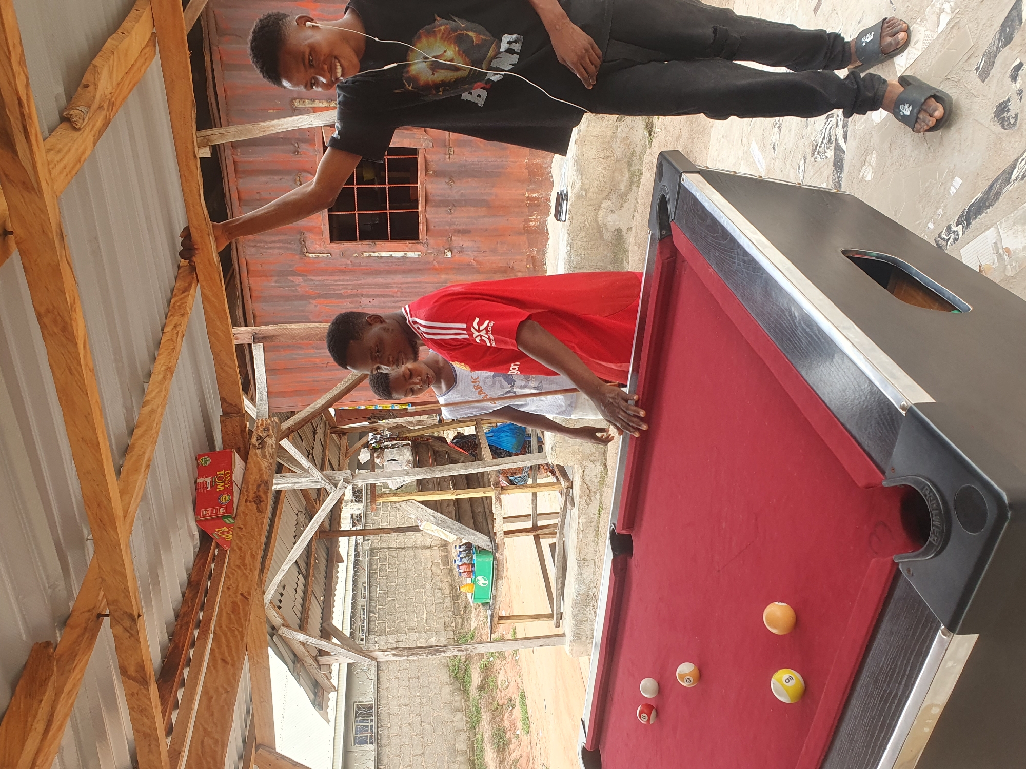 The image depicts a group of young Black males engaged in a game of pool. The setting appears to be an outdoor, open-air recreational area, possibly a local hangout or a makeshift entertainment spot. The pool table, covered in red felt, is the central focus. Several pool balls are scattered on the table, with some indicating numbers like 11, 13, and 6.
Three individuals are prominently visible. One man, wearing a black t-shirt with a graphic print and black pants, is leaning against the wooden structure of the shelter and smiling broadly, with earbuds in his ears. Another man, in a red football jersey with white stripes, is standing to the left of the smiling man, looking towards the pool table. A third individual, in a white sleeveless shirt, stands slightly behind the man in the red jersey, also observing the game.
The environment suggests a warm climate, with a corrugated metal roof supported by wooden beams providing partial shade. In the background, a weathered corrugated metal wall, possibly part of a building or container, is visible. Other elements in the background include a brick wall, some vegetation, and what appears to be a wooden shelf holding several bottles of drinks. The ground is paved with light-colored tiles with dark patterns. The overall impression is a casual, communal gathering centered around a game of pool, likely in a less formal urban or semi-urban area. The location is identified as Lokoja, Nigeria.