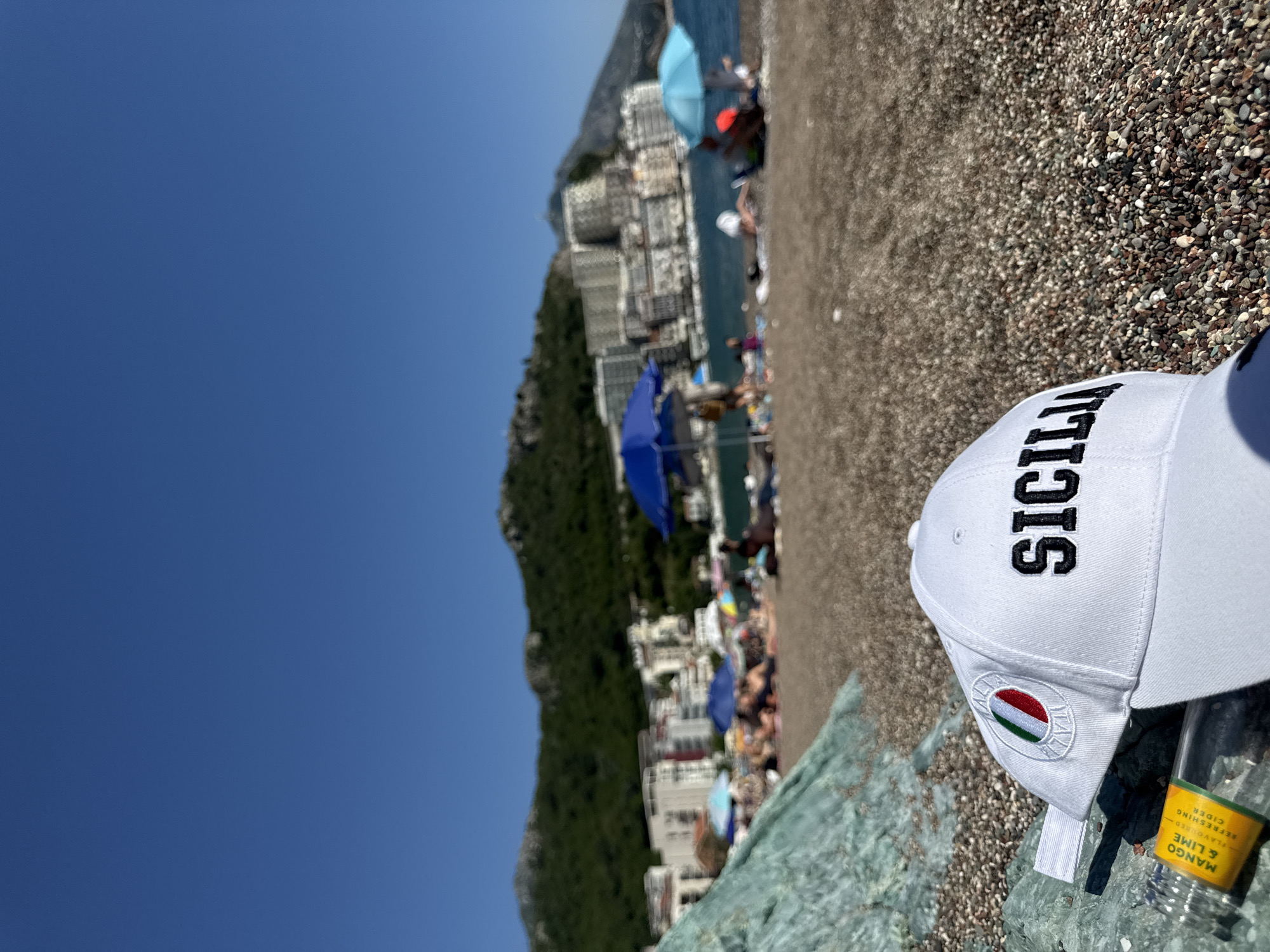 The image captures a sunny beach scene in Rafailovići, Montenegro. In the foreground, a white baseball cap, embroidered with "SICILIA" in black capital letters and featuring a circular Italian flag emblem, rests on prominent greenish-blue rocks. Adjacent to the cap is a clear bottle with a yellow label indicating "MANGO & LIME FLAVOURED REFRESHING CIDER." The ground consists of a pebbly beach with stones of varying hues. The midground shows numerous individuals relaxing under blue and light blue umbrellas on the blurred beach, with the calm sea visible beyond. The background features green, tree-covered mountains rising behind a dense array of multi-story buildings, likely hotels or residential structures, lining the coastline under a clear, cloudless blue sky.