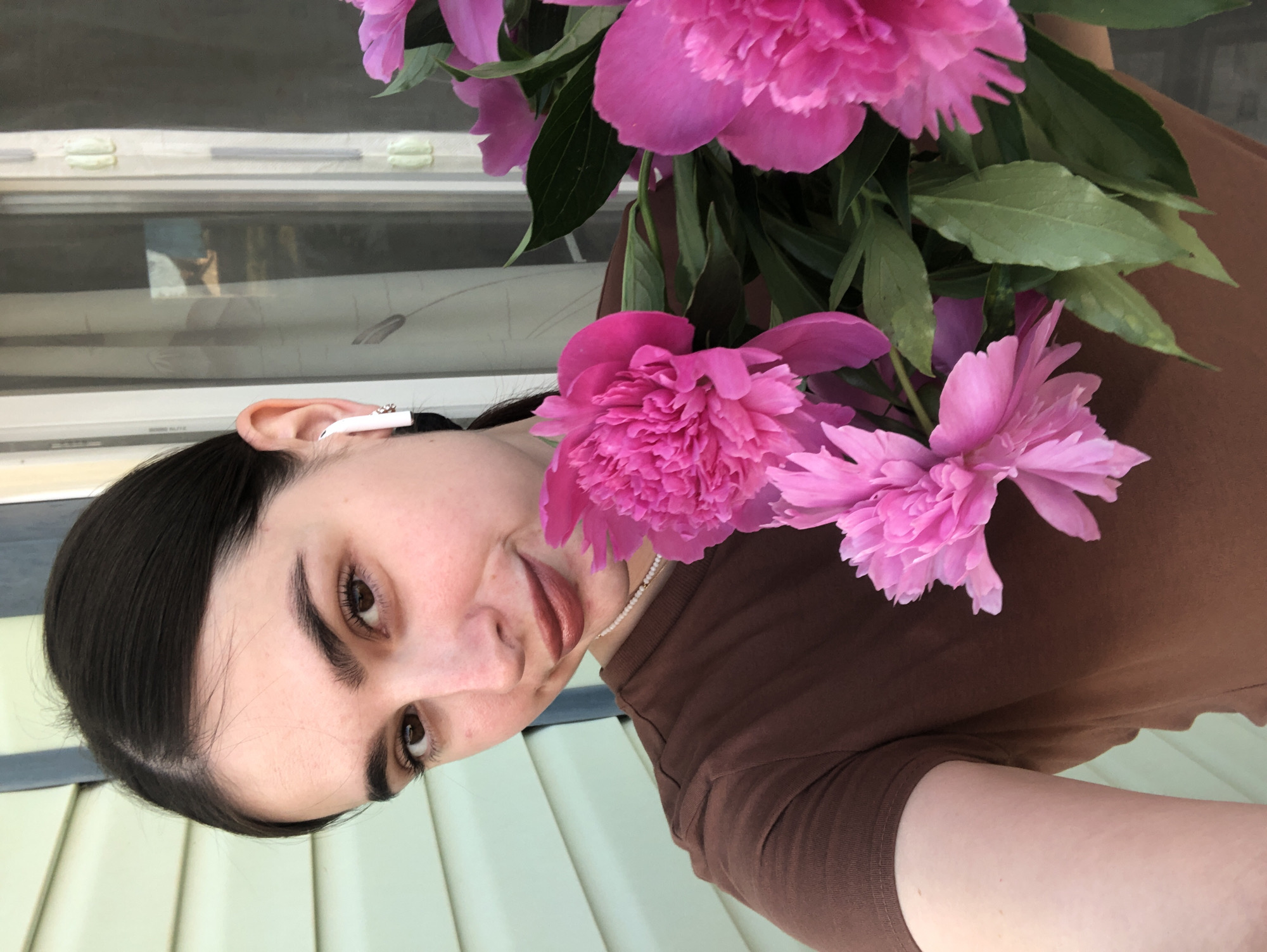 The image shows a young woman holding a bouquet of pink peonies. She is wearing a brown t-shirt and has a white earbud in her right ear. The background appears to be an exterior wall of a building, possibly a house, with light green siding. There is also a window with sheer curtains visible. The woman is looking towards the camera with a slight smile. The peonies are large and vibrant pink, with many petals. The leaves of the peonies are a deep green. The lighting suggests it is daytime, possibly late afternoon, with the sun casting a warm glow. The overall impression is a candid portrait of the woman with flowers. There is no indication of the specific location within Noviy, Russia.