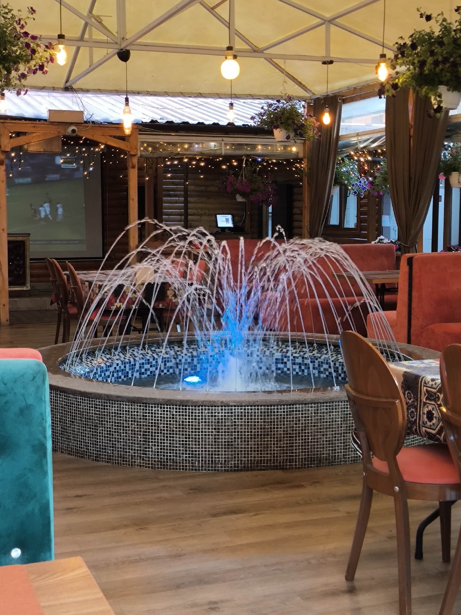 This image captures an unoccupied, semi-enclosed restaurant or cafe area, likely an outdoor patio or gazebo, located in an Unknown city, Unknown state. The scene is illuminated by artificial lighting, suggesting evening or nighttime.
Centrally positioned, a circular fountain features a basin with a mosaic tile exterior and interior lip, composed of small, square tiles in alternating light and dark grey. Multiple streams of water spray upwards, illuminated from within by a prominent blue light. The floor is constructed from light-toned wooden planks.
Seating arrangements are visible in the background. A partial teal-colored upholstered seat is present in the left foreground. Further back on the left, two wooden chairs with reddish-orange upholstered seats flank a table adorned with a patterned tablecloth. On the right, several reddish-orange upholstered booth seats and additional wooden chairs with matching seats are arranged around tables, one also featuring a patterned tablecloth.
The overhead structure includes a white metal truss system supporting a light-colored fabric or canvas roof. Multiple pendant lights, some with exposed Edison-style bulbs and others with white spherical shades, hang from this structure, casting warm yellow light. Strands of small, warm-toned string lights are draped across wooden beams and structural elements. Numerous hanging baskets containing purple and dark pink flowering plants are suspended throughout the space.
The walls are constructed from horizontal wooden logs or planks, exhibiting a rustic aesthetic. In the left background, a large white projection screen displays a live football or soccer match, showing players on a green field. A projector unit is mounted on a wooden beam above this screen. In the right background, a small desktop computer monitor with an attached keyboard rests on a wooden surface against the log wall. Brown curtains are drawn to the sides of large windows. No individuals are present in the image.