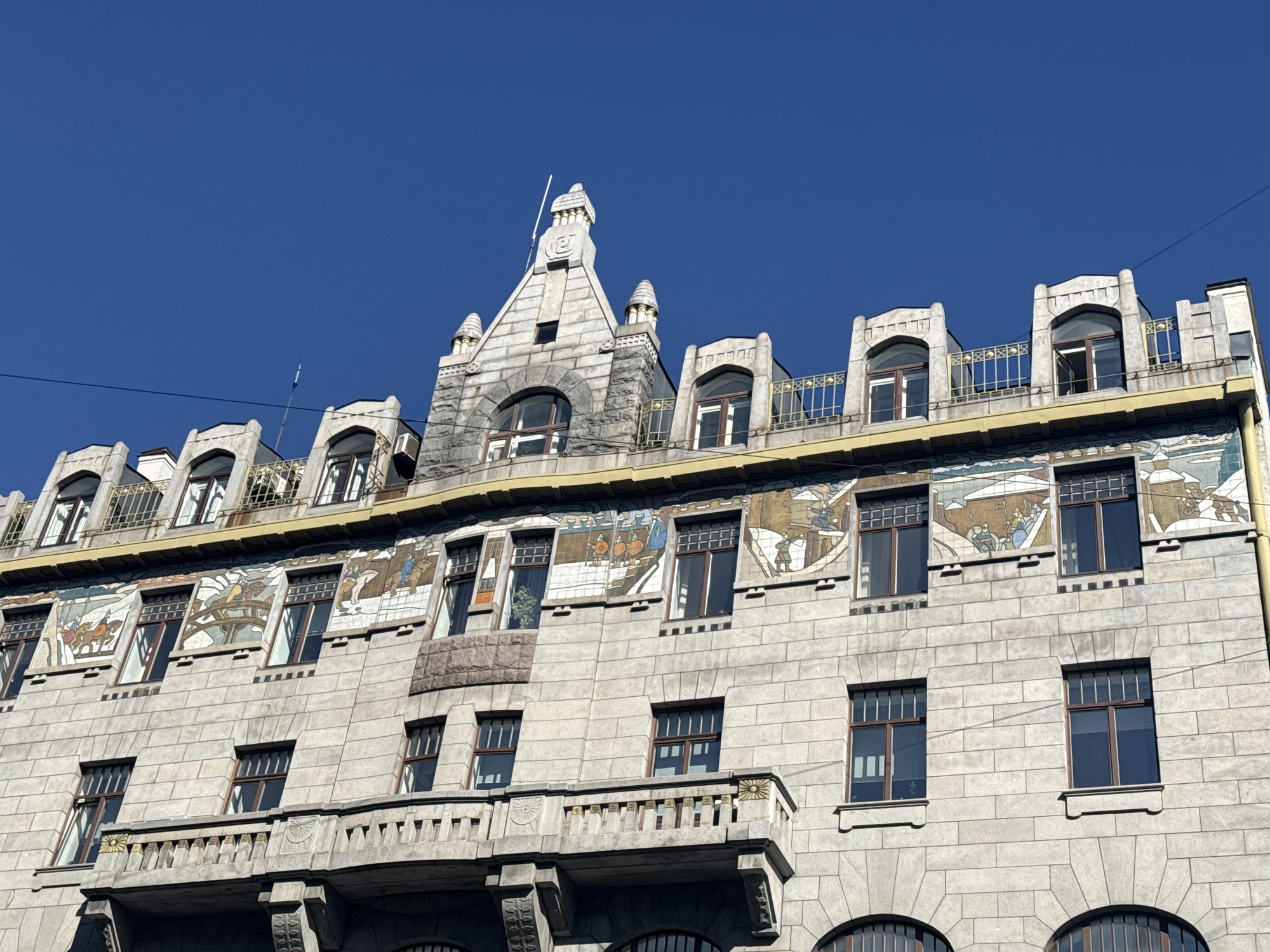 An upward-angled shot captures the elaborate facade of a historic building in Saint Petersburg, Russia, against a brilliant, cloudless blue sky. The multi-story structure is primarily clad in light-colored, rough-hewn stone blocks.
A distinctive feature is a long, colorful mosaic band positioned across the building's upper level, above a row of rectangular windows. The mosaic illustrates various scenes, primarily depicting people with horses and carriages in what appears to be a snowy, winter landscape, hinting at historical or folkloric narratives.
The building's roofline is highly ornate, featuring multiple dormer windows with decorative stone surrounds and intricate golden railings. At the very top, a prominent gabled section rises centrally, topped with a spire-like finial and flanked by smaller, conical architectural elements. Small air conditioning units are visible near some of the upper windows.
Below the mosaic, the facade continues with more rows of windows, some rectangular with dark frames, others featuring decorative grilles. A stone balcony with an ornate railing, including circular, sun-like motifs, extends from the lower visible floor. The architectural style, characterized by detailed craftsmanship and diverse textures, suggests an Art Nouveau influence. The scene is bathed in bright daylight, casting clear shadows and highlighting the building's intricate details. No visible text is present.