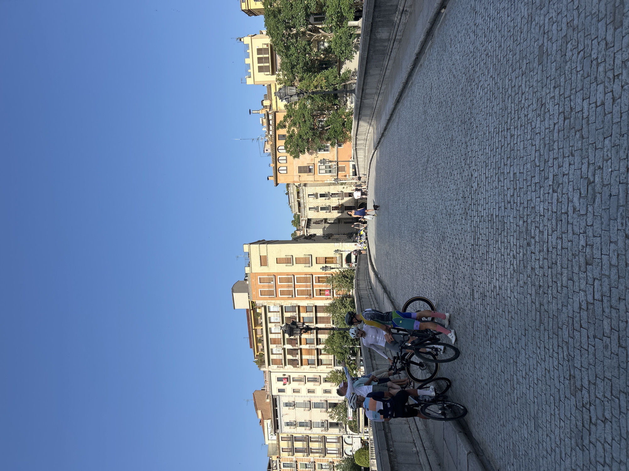 A group of cyclists are standing on a cobblestone bridge in Girona, Spain. The bridge is lined with stone barriers and lampposts. In the background, several buildings with colorful facades and balconies are visible. The sky is clear and blue. Some people are walking on the bridge in the distance.