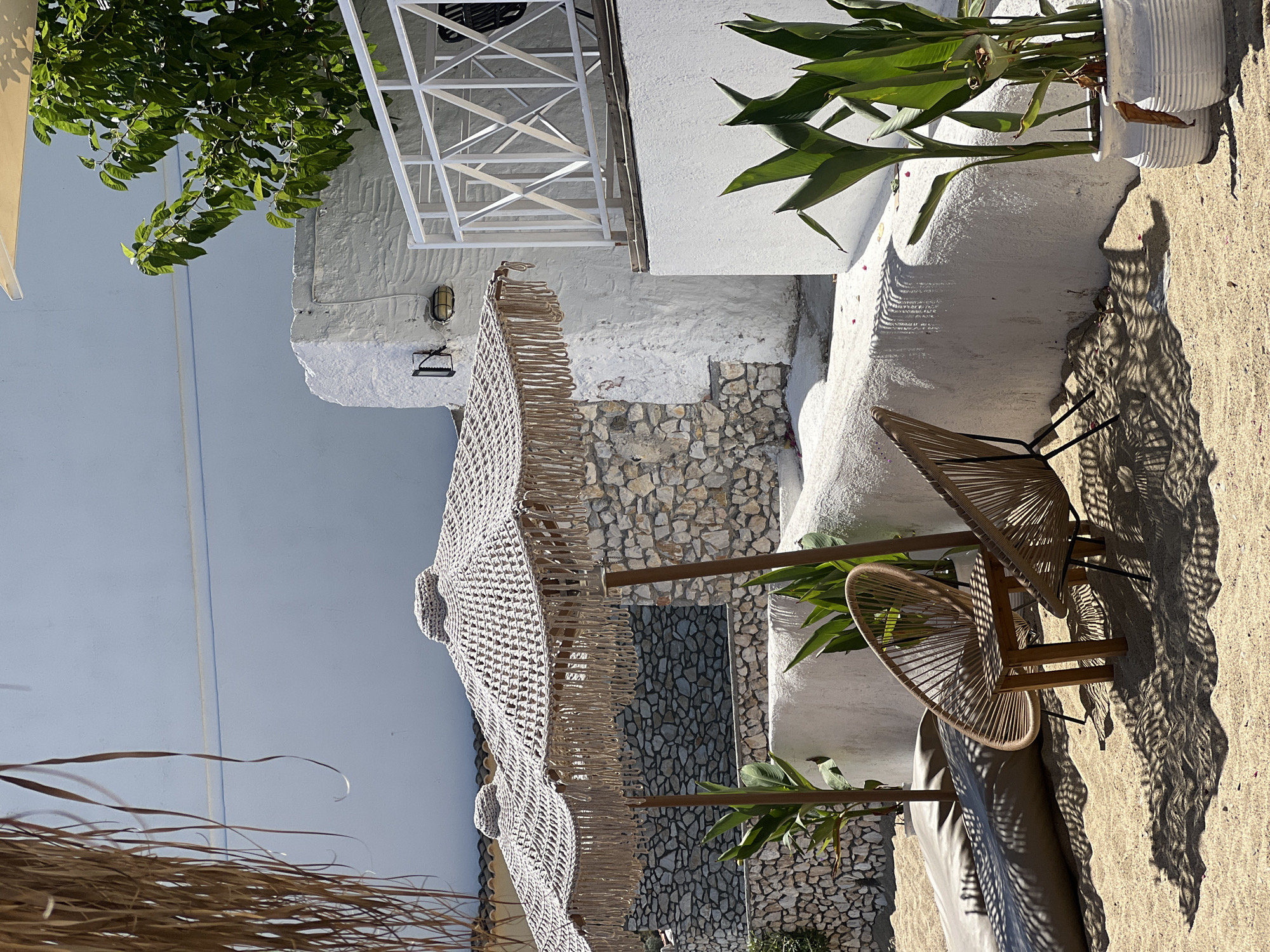 This image captures an outdoor seating area on a sandy surface, likely a beach or a beachfront establishment. The scene is bathed in bright sunlight, casting distinct shadows from the objects onto the sand. In the foreground, two woven chairs and a small wooden table are arranged. Behind them, two large, macrame-style umbrellas provide shade. The backdrop features whitewashed walls and a stone wall, suggesting a Mediterranean architectural style. Green plants in white pots are placed along the wall, adding a touch of natural color. A white railing runs along a raised platform in the background. The overall ambiance is one of a serene, sunny day in a coastal location. This location is identified as Achladias, Greece.
