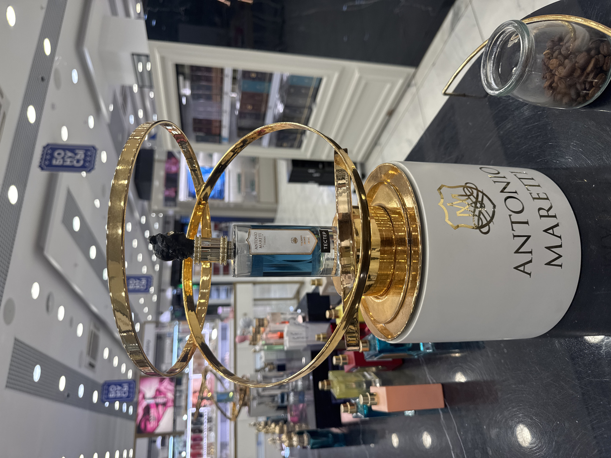 A perfume display featuring a bottle of "ANTONIO MARETTI FIRENZE" in blue liquid is presented on a white cylindrical base with gold accents. The display is ornate, with gold rings encircling the perfume bottle. In the background, shelves are stocked with various perfume bottles and products, suggesting a retail environment, possibly a department store or a specialized fragrance shop. A small glass jar filled with coffee beans sits to the right of the display base. The lighting is bright, with overhead lights visible, and the overall impression is one of luxury and product presentation. The image was taken in a location within the city of Yekaterinburg, Russia.