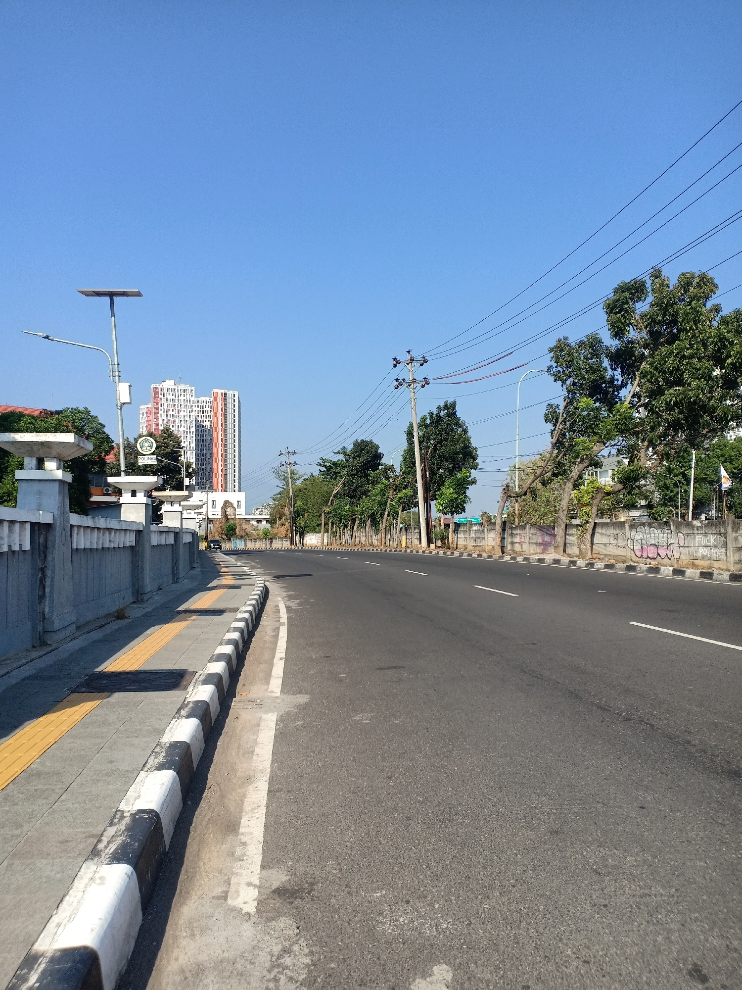This is a low-angle, eye-level, full shot of a street in Semarang, Indonesia. The street is paved with asphalt and has white lane markings. On the left side of the street, there is a sidewalk with tactile paving and a curb painted with black and white stripes. A concrete barrier separates the sidewalk from the road, topped with white pillars. A street lamp with a solar panel is visible above the barrier. In the background, there are several tall buildings, some with colorful facades, and a cluster of trees along the right side of the road, with power lines running above them. There is no visible human presence or activity. The sky is clear and blue.