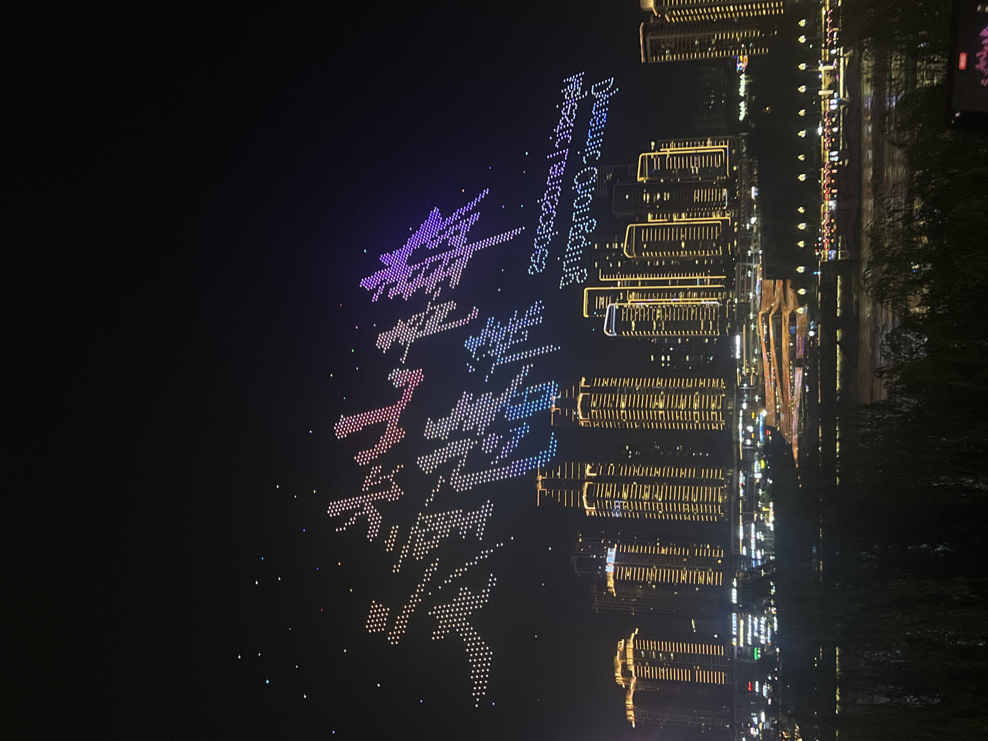 A nighttime drone light show is taking place above a cityscape featuring illuminated skyscrapers and a river. The drones are arranged to form large Chinese characters that appear to say "壮丽山河" (magnificent mountains and rivers) and "重庆" (Chongqing). Below the drone display, the buildings are lit up with yellow and white lights, and the river reflects the city lights. A portion of the text "Fantastic Chongqing" is also visible, spelled out in English by the drones, but it is inverted. The scene is located in the Jiangbeicheng Street (江北城街道) area of Chongqing, China.