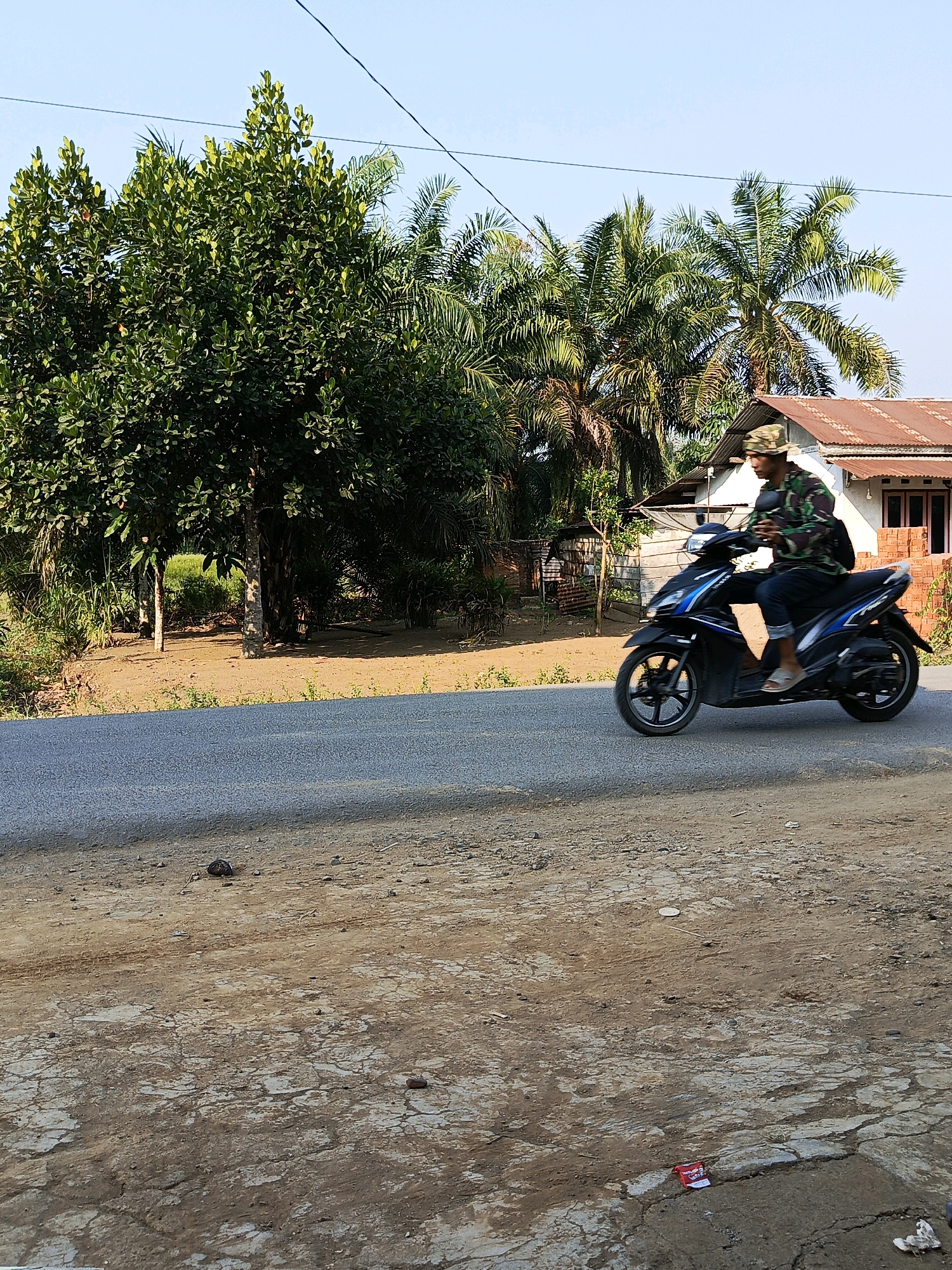 A person wearing a camouflage hat and jacket rides a dark blue and black motorcycle on a paved road. The rider is seen from the side, with their left leg extended forward and their right leg on the footrest. The motorcycle has blue accents and black wheels. In the background, there are several palm trees and a building with a reddish-brown roof. A large green tree with dense foliage is visible to the left of the motorcycle. The ground beside the road is dry and appears to be dirt with some cracks. The sky is clear and blue. The scene depicts a rural or semi-rural environment in Megang Sakti I, Indonesia, possibly during the day.