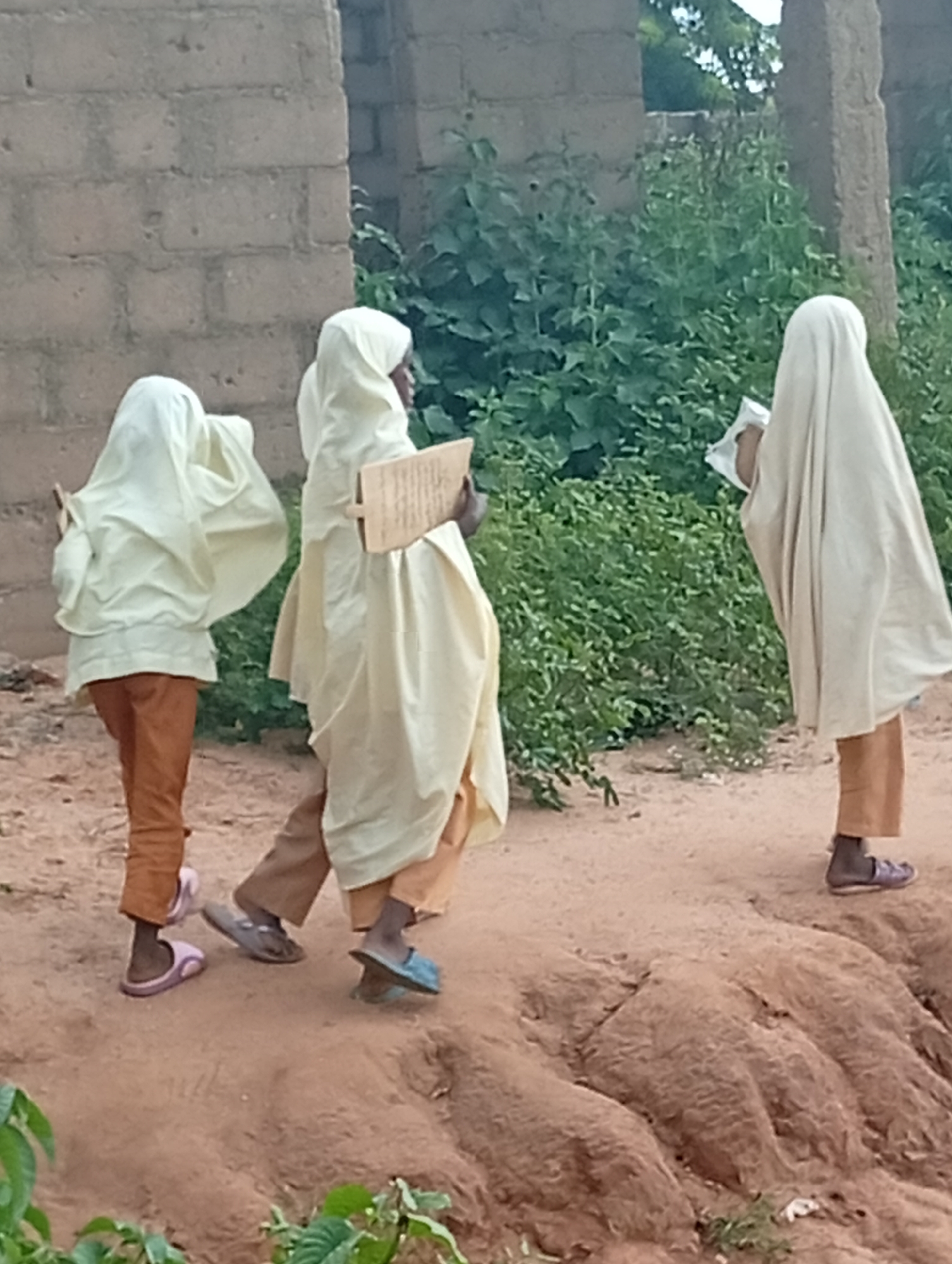In Tumfure, Nigeria, three individuals, likely children, are walking on a dirt path. The two in the foreground are wearing light-colored robes and head coverings. The person on the left is wearing orange pants and pink slippers. The middle person is wearing brown pants and dark sandals. They appear to be carrying educational materials, with the middle individual holding a wooden board with writing. The third individual, on the right, is also wearing a light-colored robe and head covering, brown pants, and dark sandals, and is holding a white object. They are walking away from a rough brick wall and a dense green foliage. The ground is uneven and sandy, with some small plants visible.
