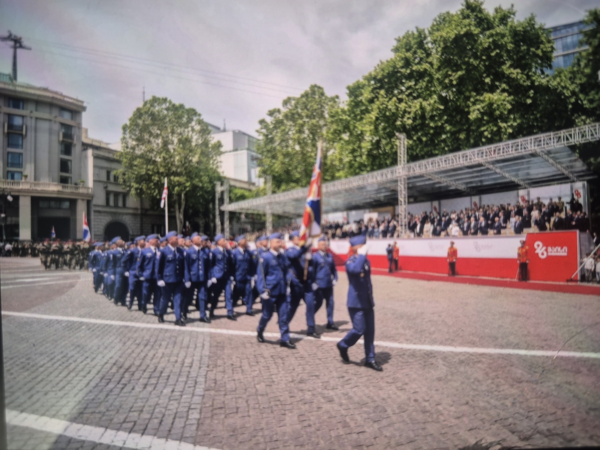 The image depicts a military parade in Tbilisi, Georgia. A column of soldiers in blue uniforms marches on a cobblestone square, with spectators observing from a grandstand in the background. The Georgian flag is visible. The architecture suggests a city center location. The event appears to be a formal ceremony. The environment is daytime with overcast skies.