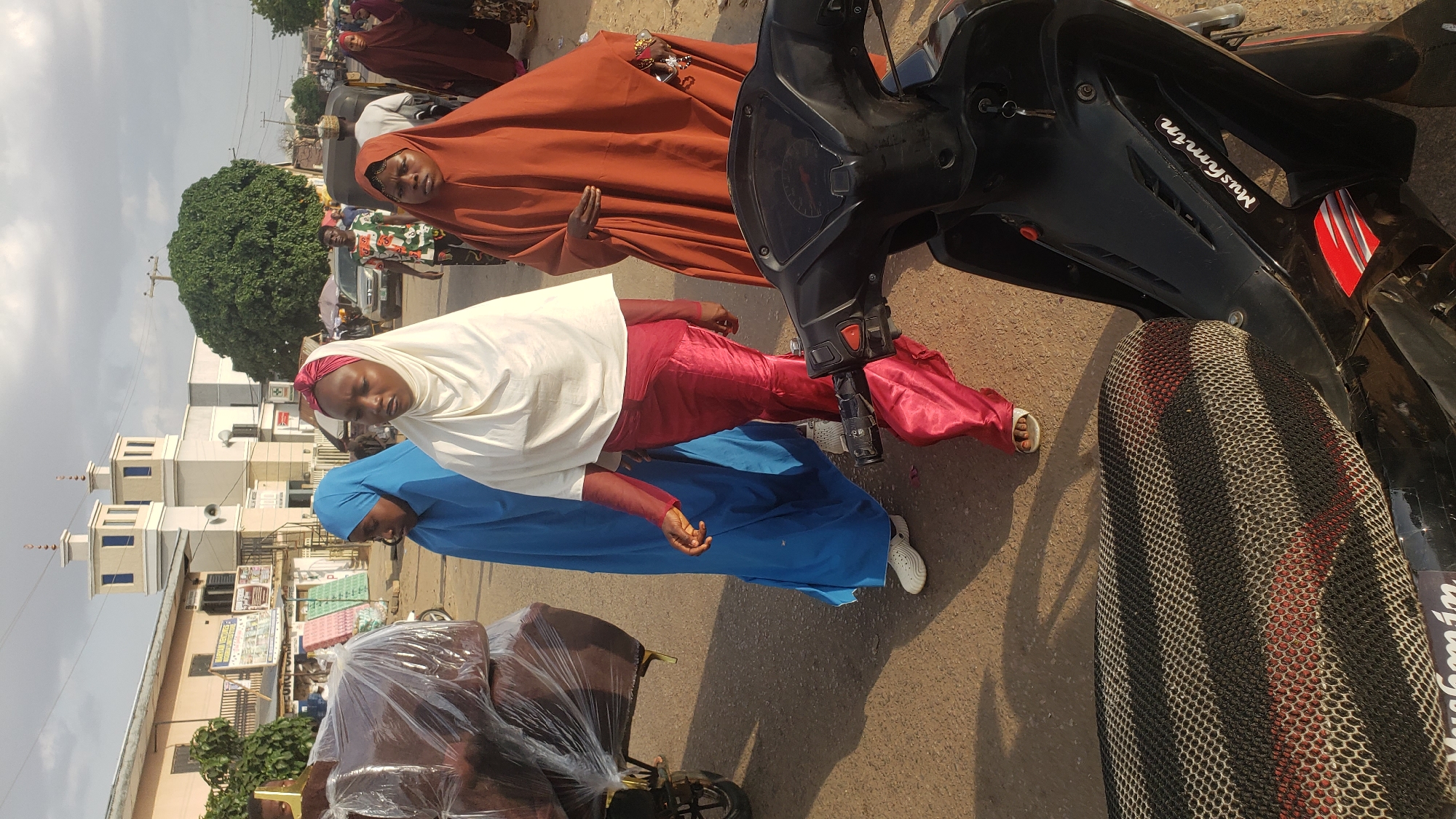 The photograph, taken in Kawo, Nigeria, shows three women dressed in traditional Muslim attire. The woman closest to the camera is wearing a blue hijab and is seated near a pushcart loaded with furniture wrapped in plastic. Adjacent to her is another woman in a white hijab and a red garment. Further back, the third woman is wearing a brown hijab. They stand on an asphalt road. To the right is a black motorcycle with the inscription "Mushmin" visible. In the background, buildings and trees are visible, including what appears to be a mosque with distinct towers. People are blurred in the distance.