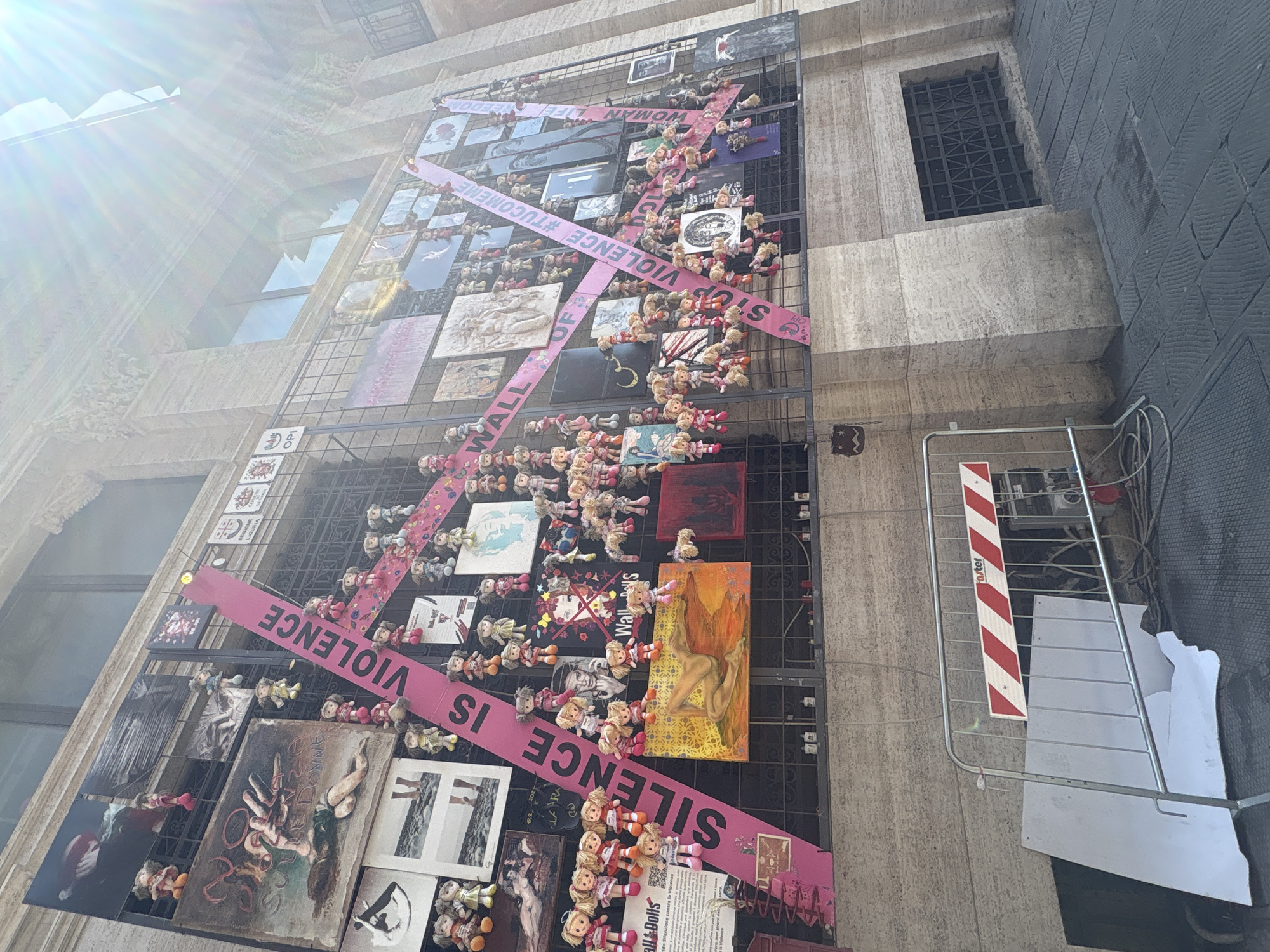 Here is a description of the image:
The image depicts a display titled "Wall of Silence is Violence" erected on the exterior of a building, possibly in Genoa, Italy, based on the Liguria sign present. The display is a wire grid filled with various artworks, photographs, and numerous small dolls, arranged haphazardly. A broad pink ribbon diagonally crosses the structure, bearing the text "SILENCE IS VIOLENCE" and other text in Italian. The display aims to raise awareness against violence, particularly against women, as indicated by hashtags and thematic elements.
The building's façade, made of light-colored stone or concrete, features a window with a dark grill. To the right of the display, a scaffold-like structure is present, with red and white striped caution markers. The overall atmosphere suggests an urban setting with a focus on social activism and artistic expression.