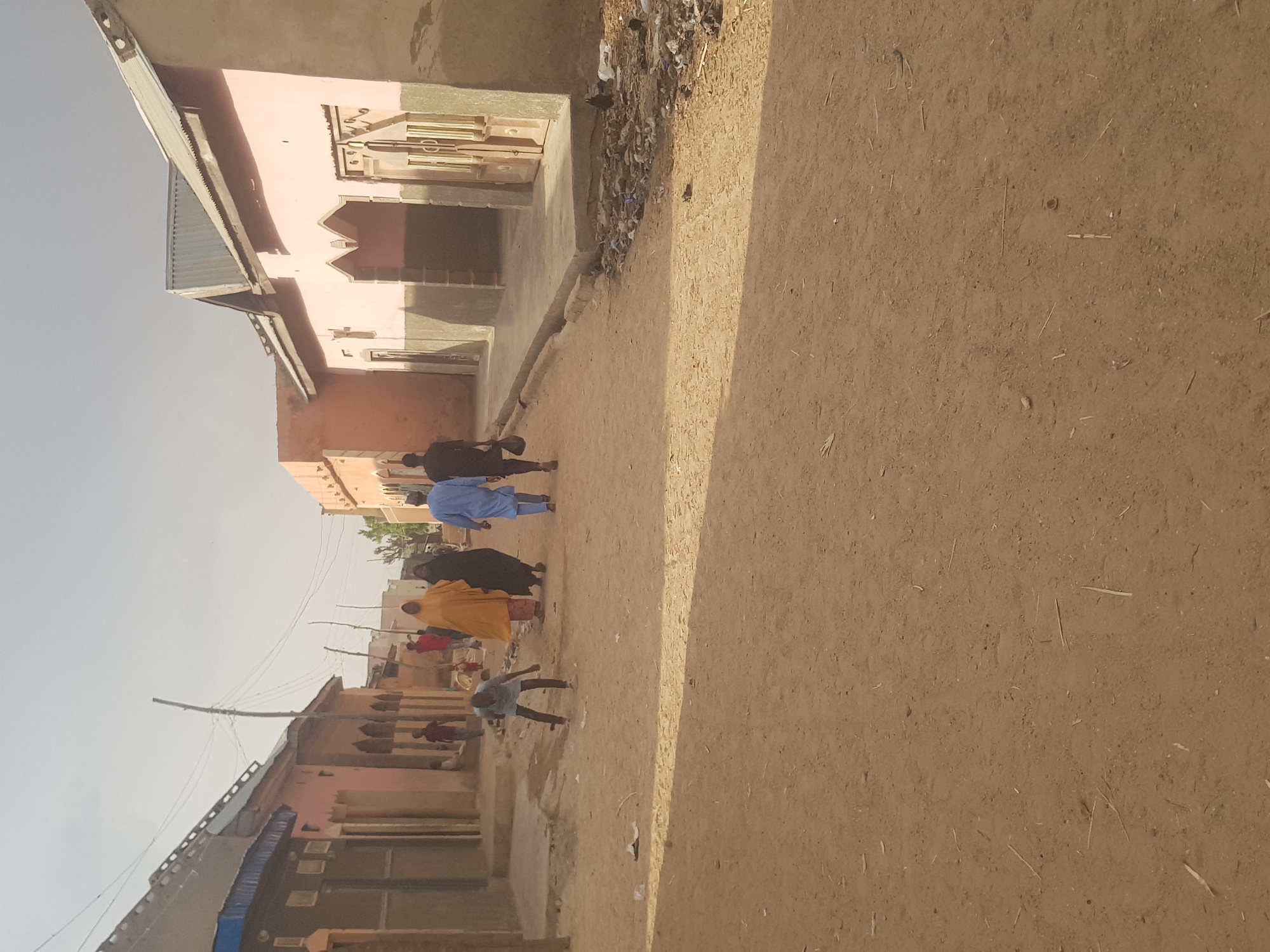 The image depicts a street scene in Dutsinma B, Nigeria. A group of people, dressed in various attires including blue and yellow robes, are walking down a dirt road lined with buildings made of light-colored materials. The buildings have simple architectural designs. The road surface is dusty and uneven. Power lines are visible overhead, connecting to buildings along the street. The sky is a pale blue, suggesting daylight. A person is seen in the middle ground bending over near the street. The scene conveys a sense of everyday life in a rural or less developed urban area.