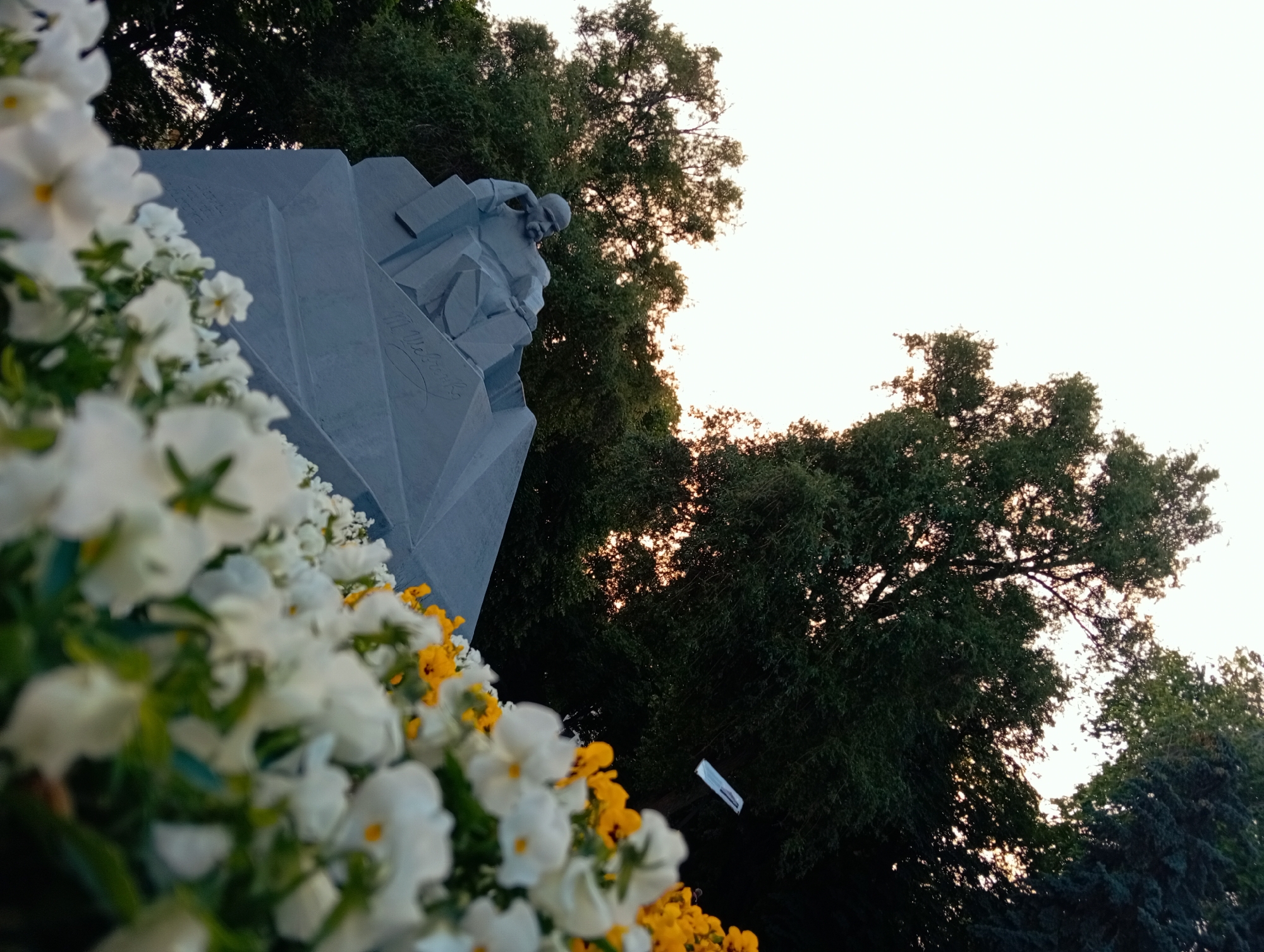 This is a low-angle, full-frame photograph of a monument in Poltava, Ukraine, with foreground floral elements. The monument is a large, abstract, light gray structure with geometric shapes, resembling carved stone or concrete. A sculpted figure, appearing to be a bald man with a beard, is depicted in a thoughtful or contemplative pose, leaning on the monument. The monument has visible inscriptions on its surface, including what appears to be a signature.
In the foreground, a dense arrangement of white and yellow flowers, likely petunias or similar bedding plants, is out of focus, creating a soft, blurred effect. These flowers extend across the bottom of the frame.
Behind the monument and flowers, a canopy of lush green trees with dark foliage dominates the midground and background. The sky is a pale, washed-out white or light yellow, suggesting it is either early morning or late evening. The lighting is soft, with some areas of the trees backlit by the sky, creating a diffused glow. The overall impression is one of a memorial or public garden space in Poltava.
