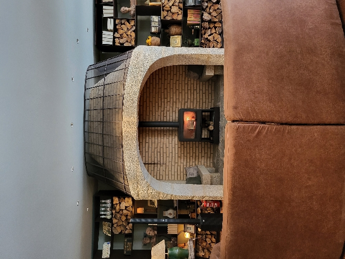 The scene depicts an interior living space in São Pedro do Sul, Portugal, centered around a large fireplace structure. A prominent, light-colored rough stone arch frames a recess lined with light brown bricks. Within this recess, a black, freestanding wood-burning stove displays glowing embers and flames behind a glass door, with additional logs stored beneath it. Two simple stone benches are positioned within the arch, flanking the stove. Above the stone arch, an inverted conical metal hood with a gridded surface extends towards the ceiling.
On either side of the central fireplace structure, dark-colored shelving units are integrated into the wall. These shelves contain numerous stacks of cut firewood, alongside assorted decorative items including books, ceramic busts, lamps, and various vessels. A black floor-standing speaker and a red fire extinguisher are visible to the left of the fireplace arch. In the foreground, the back of a large, curved, two-section brown upholstered sofa occupies the lower portion of the image. The ceiling above is light-colored and features several recessed lights. No individuals are present.